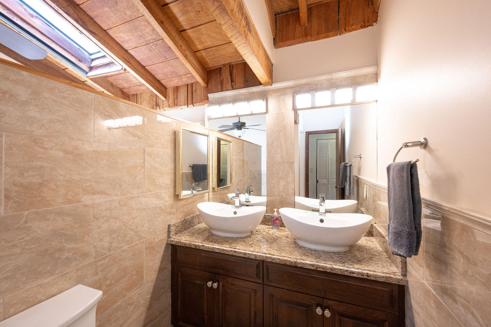 Bathroom with double vessel sinks beneath a skylight and exposed wood beams.