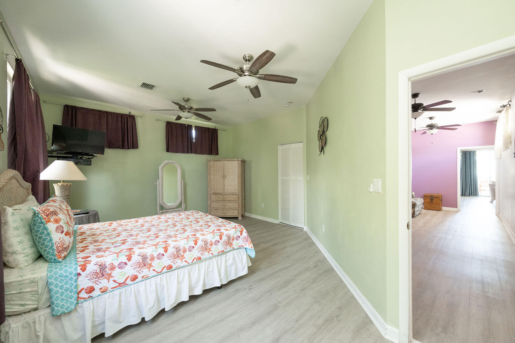 Furnished bedroom with pale green walls, coral-patterned bedding, and a doorway to a purple room.