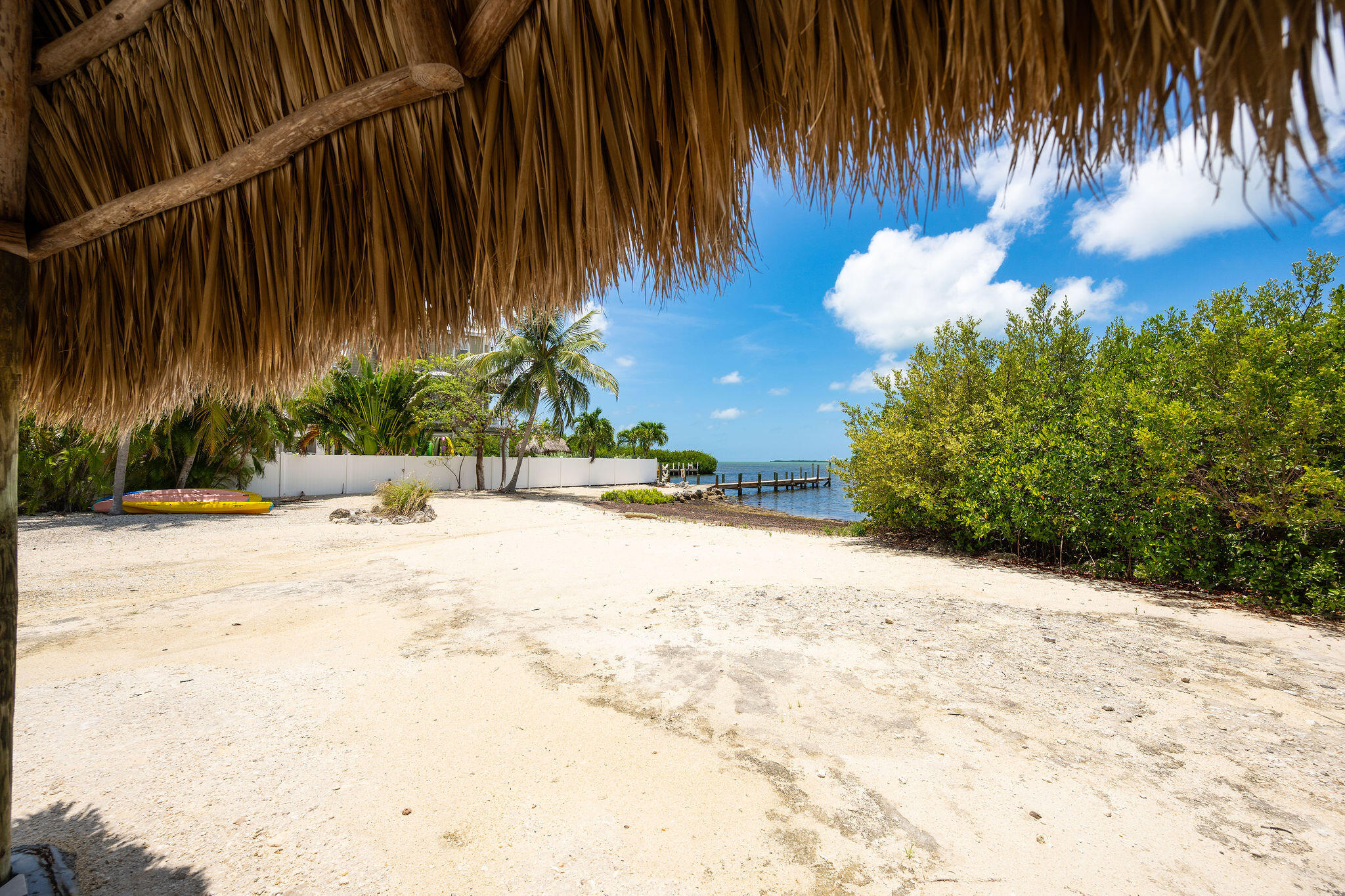 Sandy waterfront yard beneath a thatched roof, with palms, a kayak, and a dock.