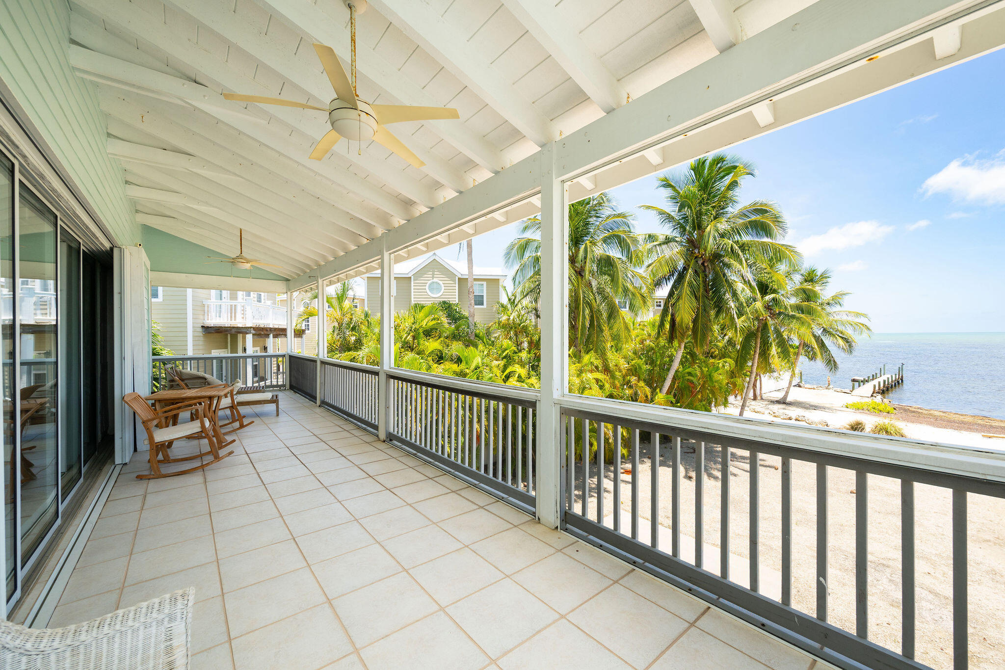 Covered tiled porch with rocking chairs overlooks palm trees, a small dock, and open water.