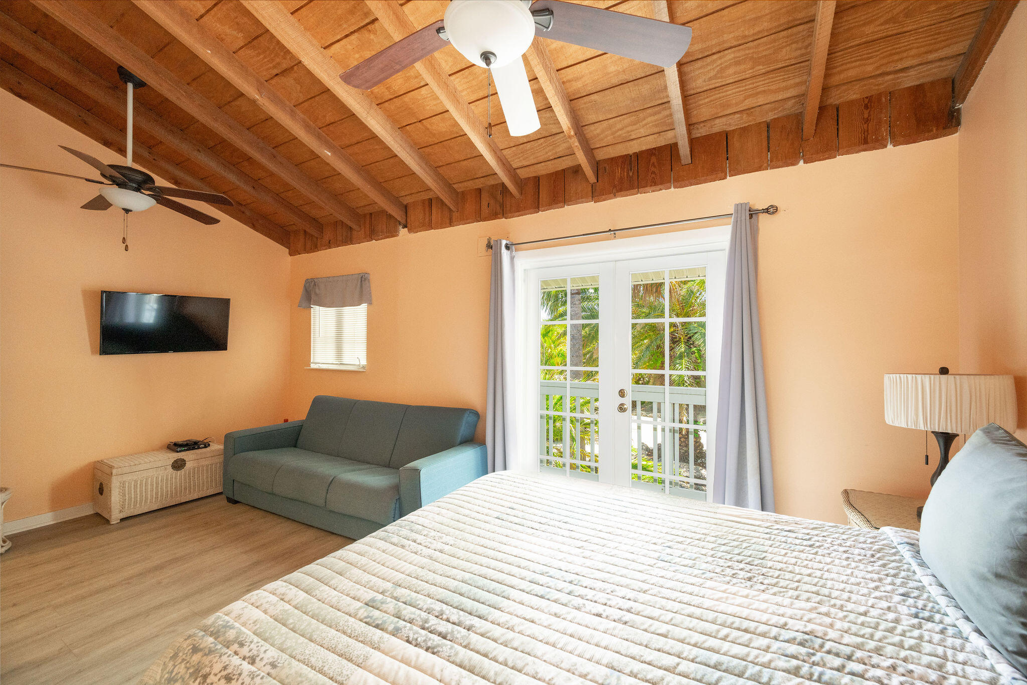Bedroom with exposed wood vaulted ceiling, gray sofa, and French doors opening to palm greenery.