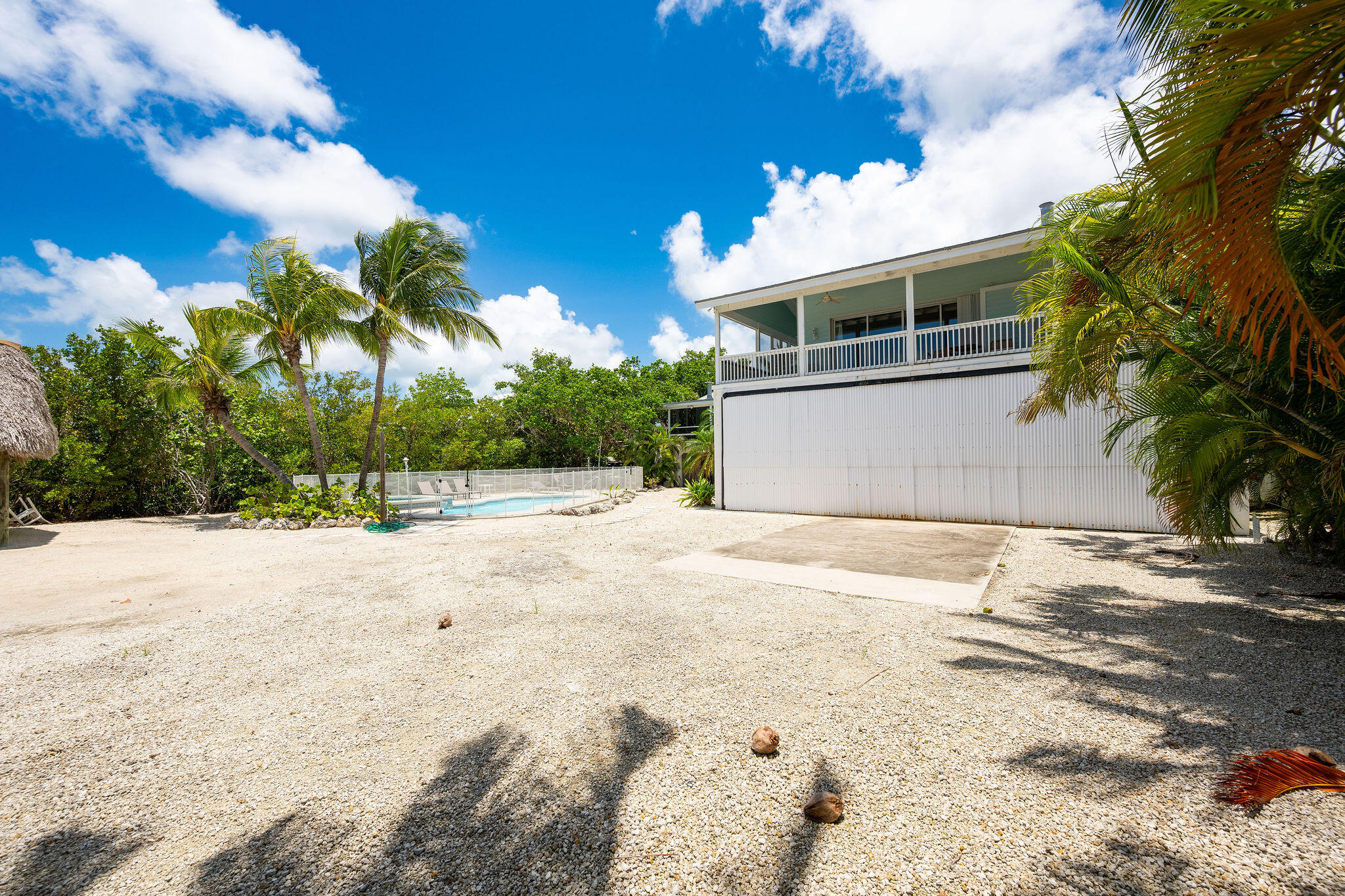 Raised coastal home with an upper balcony, fenced pool, palm trees, and a broad gravel yard.