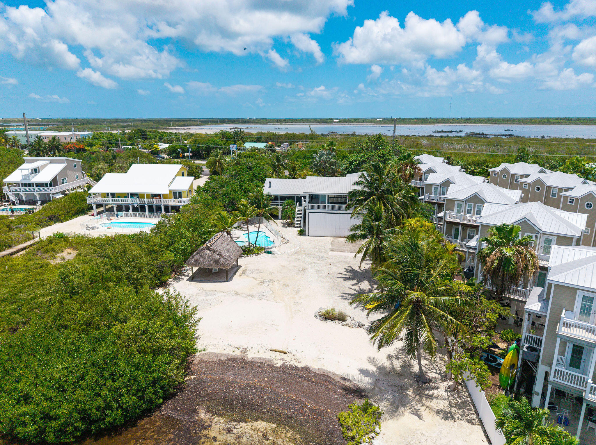 Aerial view of a sandy waterfront property with a pool, tiki hut, and neighboring homes.