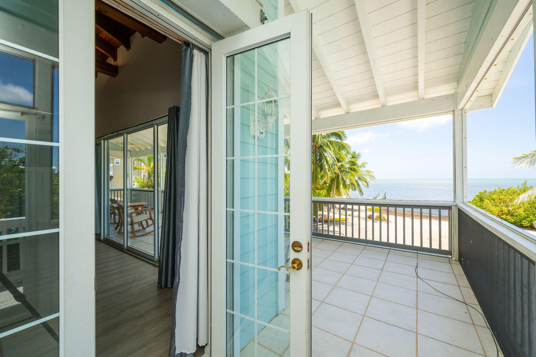 Covered balcony overlooking palm trees, shoreline, and ocean, with an open glass door.