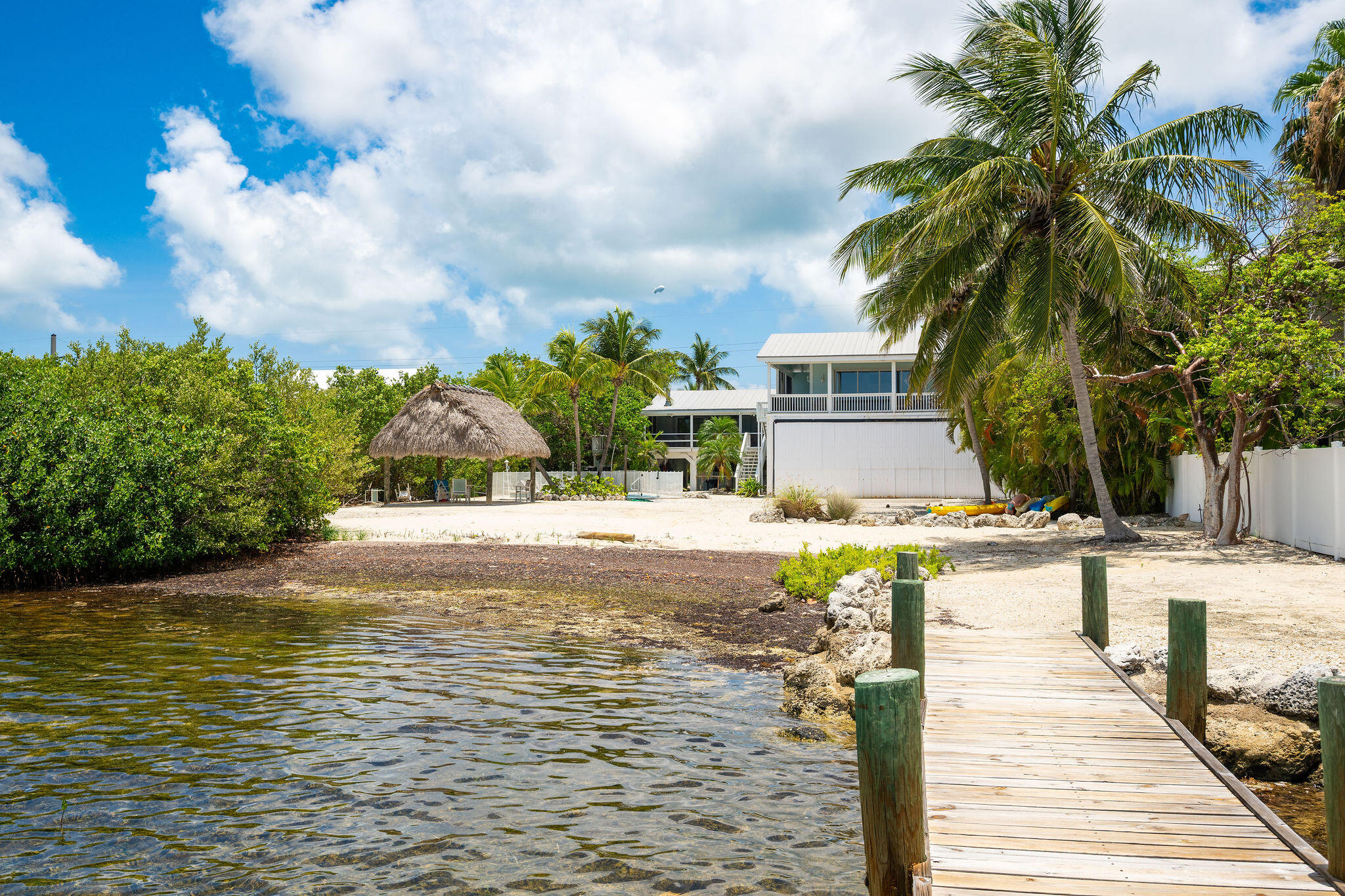 Waterfront house with a sandy palm-lined yard, thatched shelter, and wooden dock.