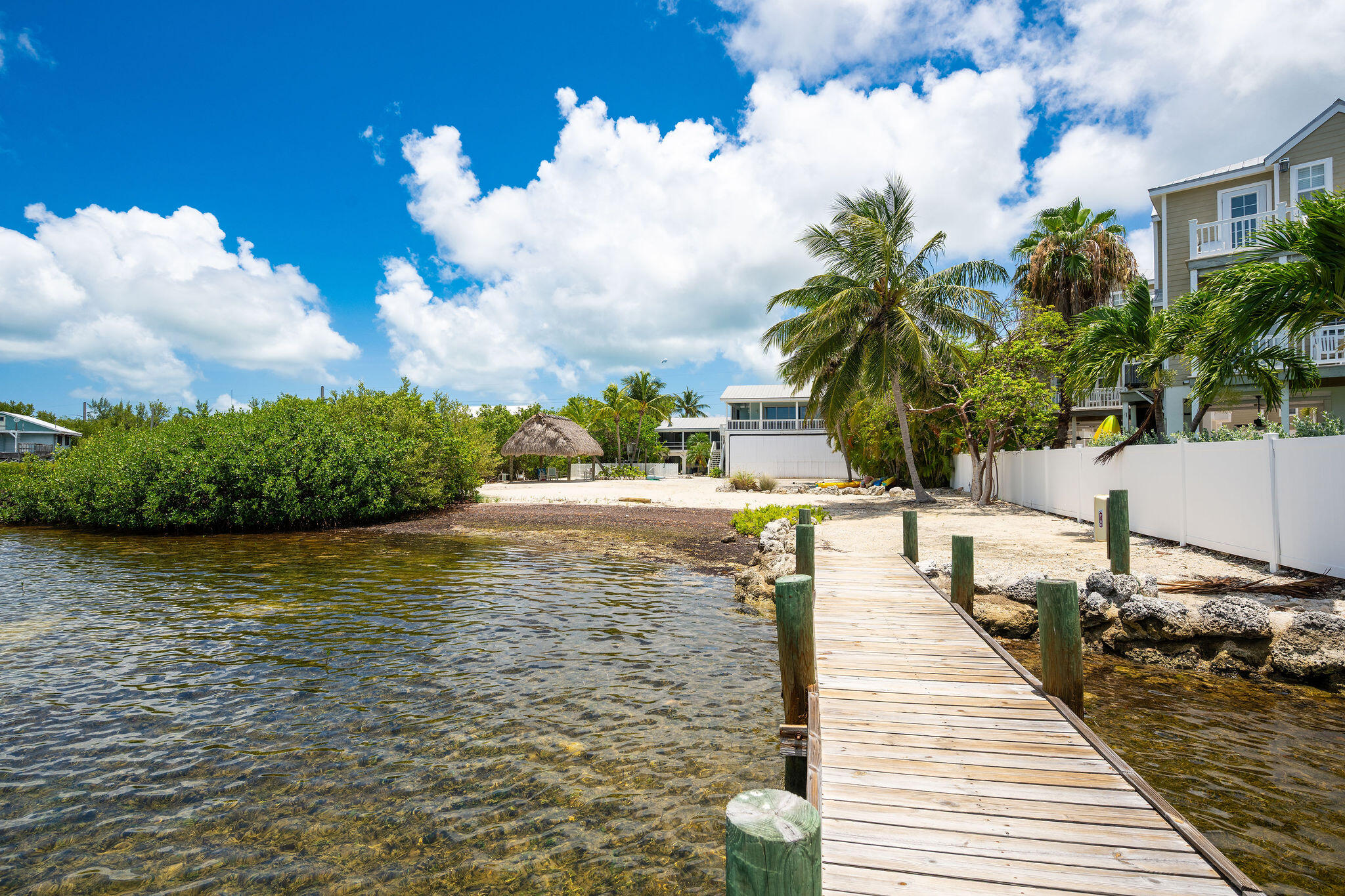 Waterfront property with a wooden dock, clear shallow water, sandy shore, and palm trees.