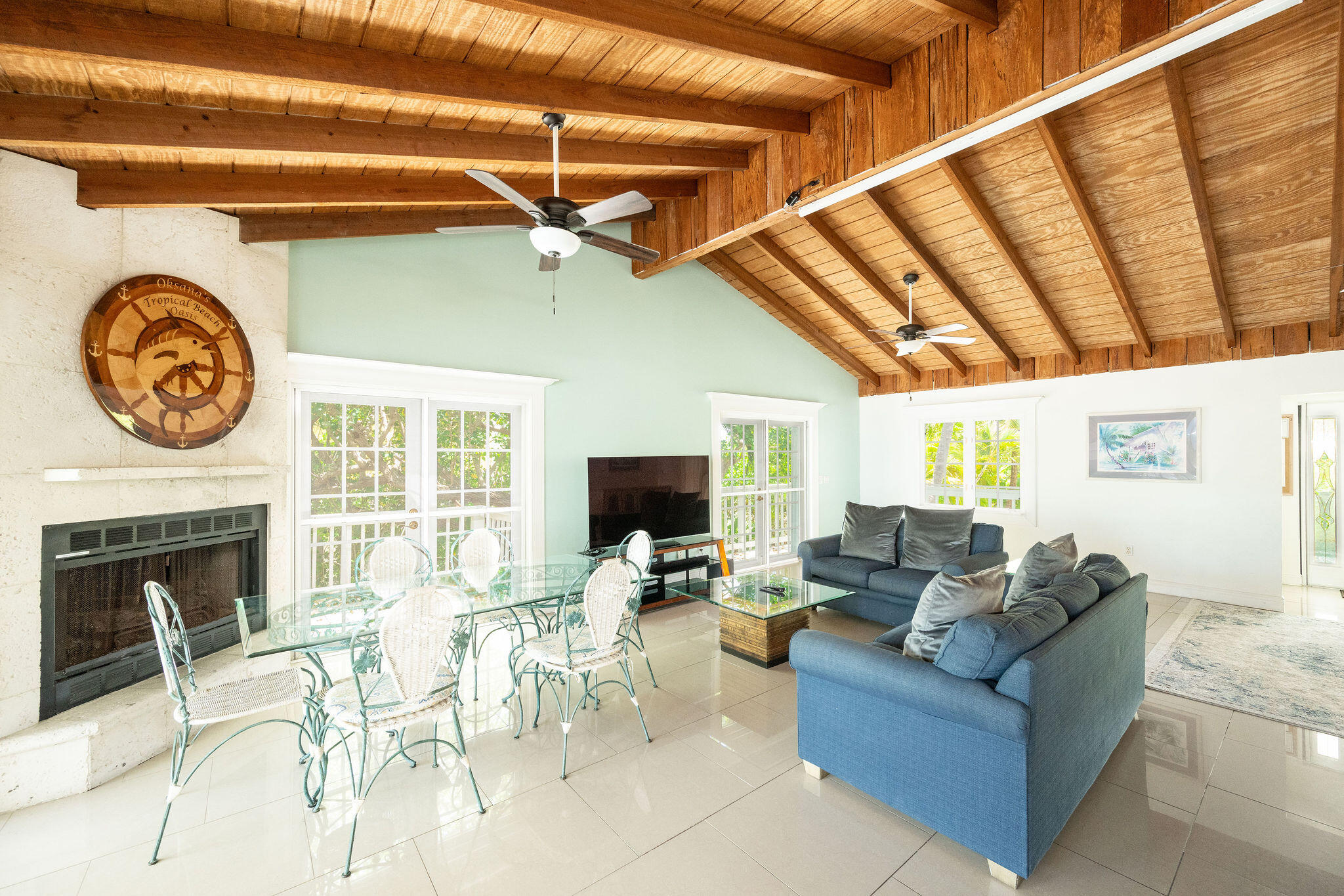 Living room with exposed wood ceiling, stone fireplace, glass dining table, and blue sofas.