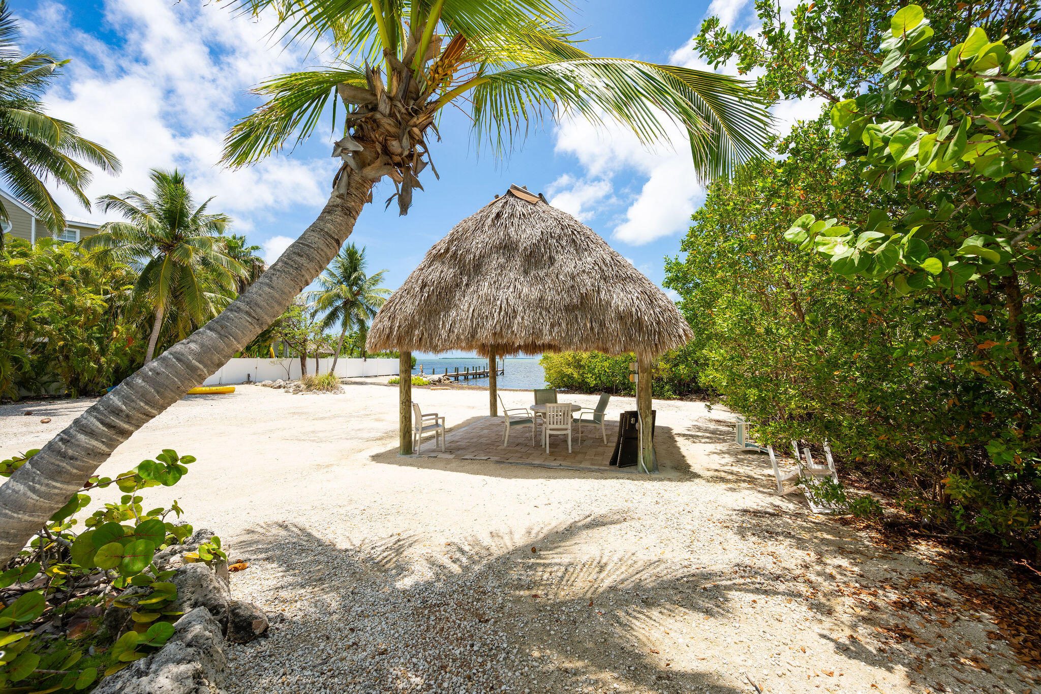 Thatched tiki pavilion with patio seating on a sandy, palm-lined waterfront.