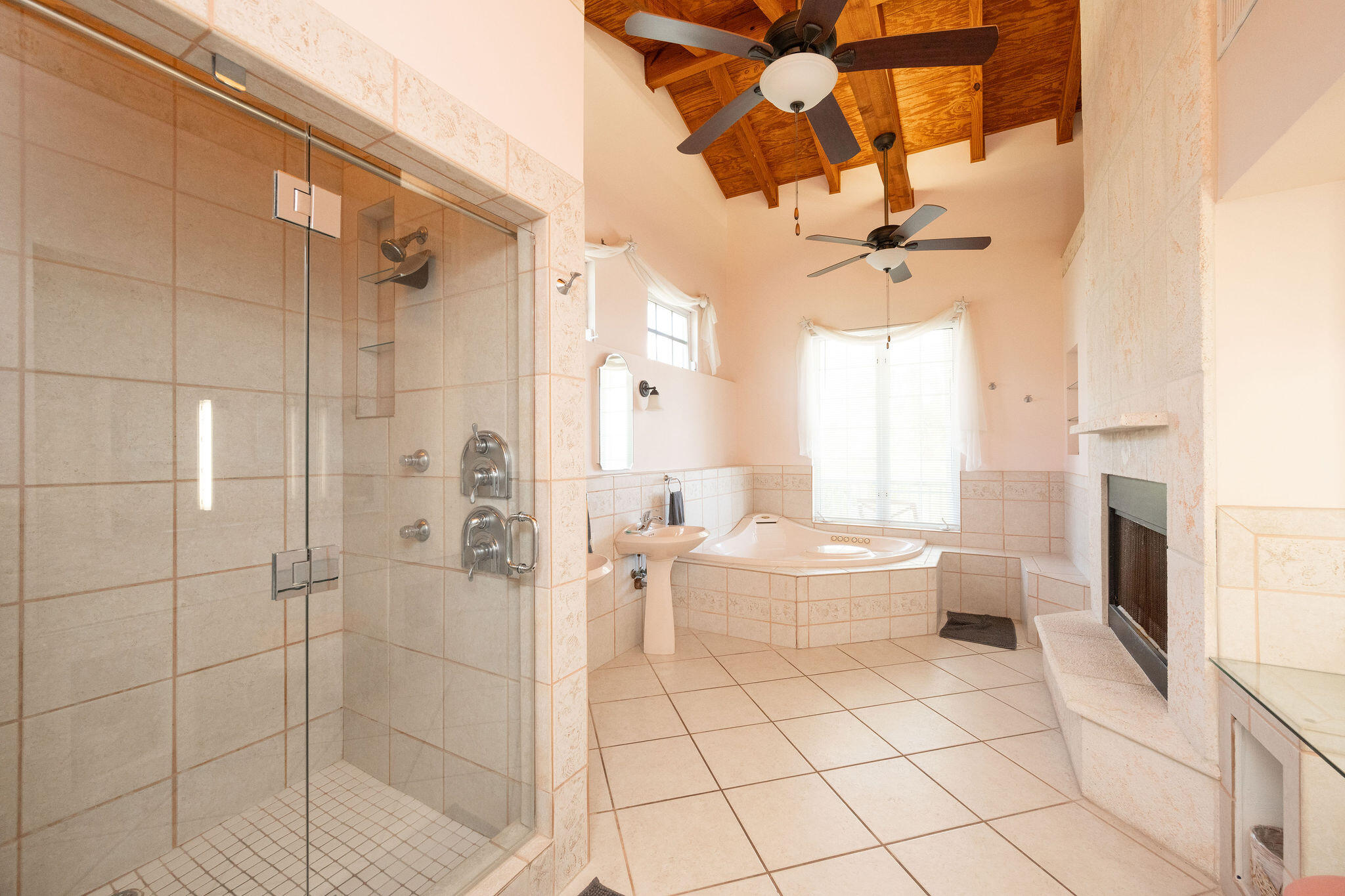 Spacious tiled bathroom with glass shower, corner soaking tub, fireplace, and vaulted wood ceiling.