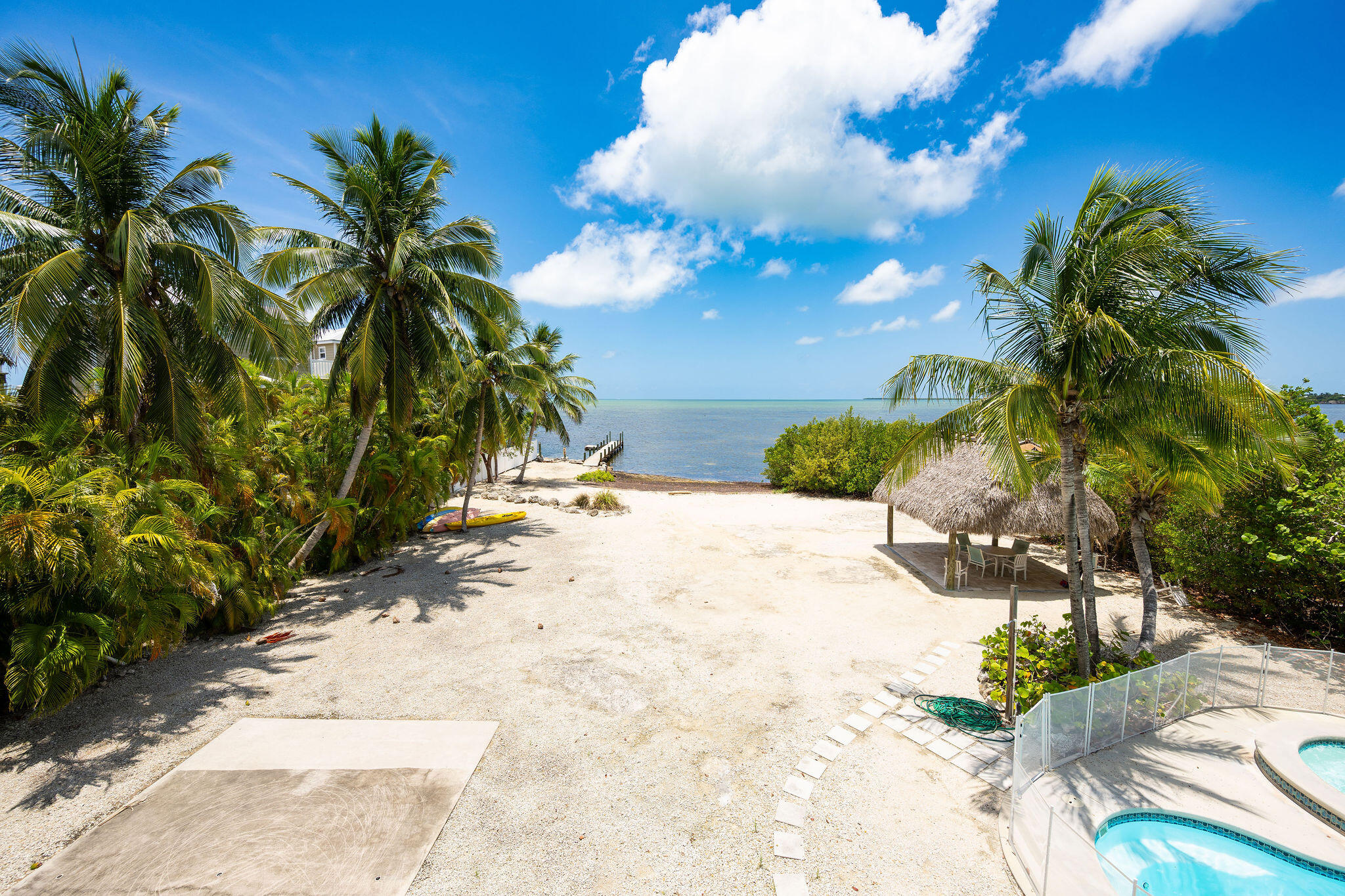 Sandy waterfront yard with palm trees, a thatched gazebo, dock, and swimming pool.