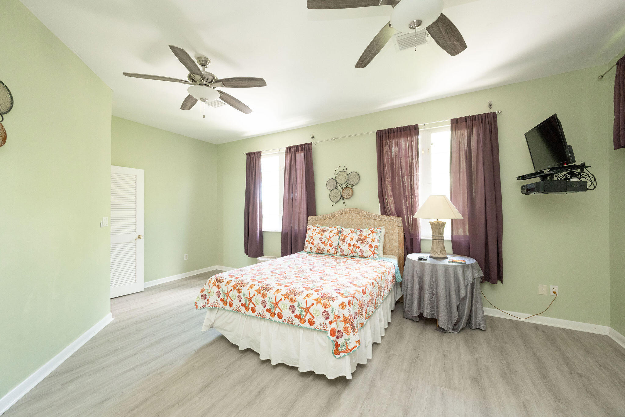 Bedroom with seashell-patterned bedding, pale green walls, purple curtains, and two ceiling fans.