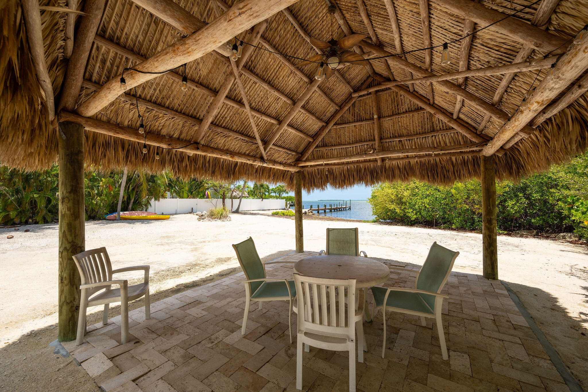 Waterfront patio under a thatched tiki pavilion, with outdoor dining furniture and a dock beyond the sandy yard.