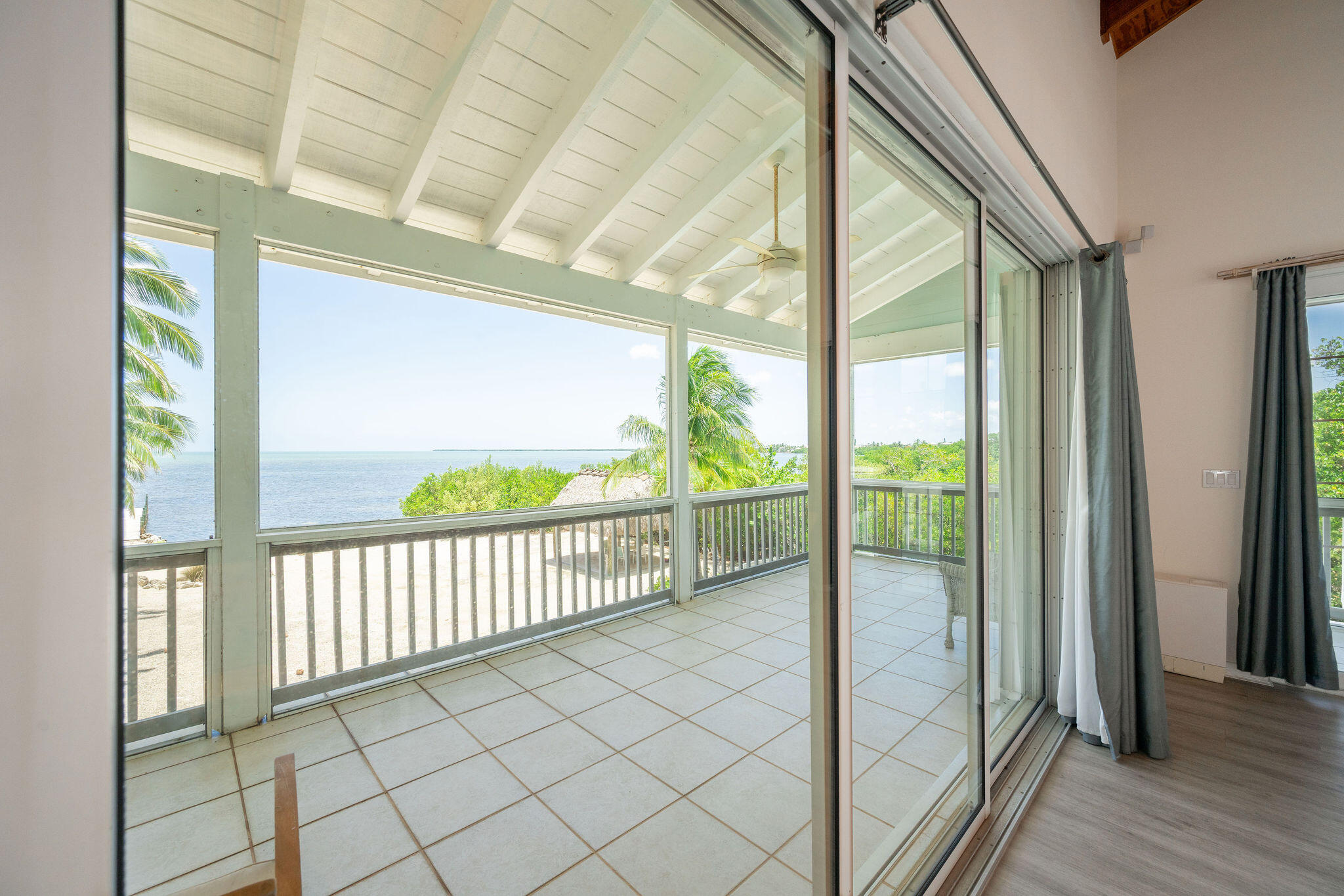Sliding glass doors open to a covered tiled balcony overlooking palm trees and open water.