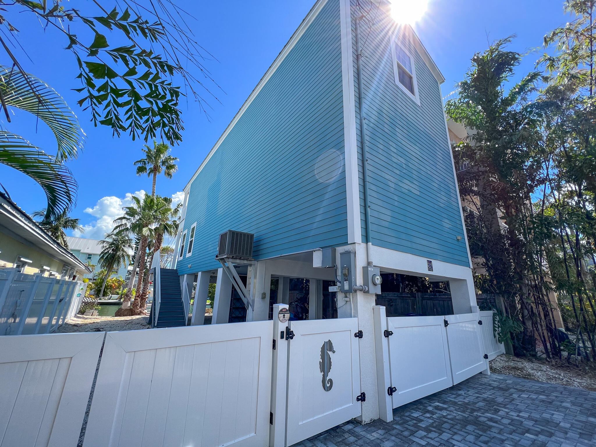 Elevated blue house with white fencing, a paved driveway, and palm trees.