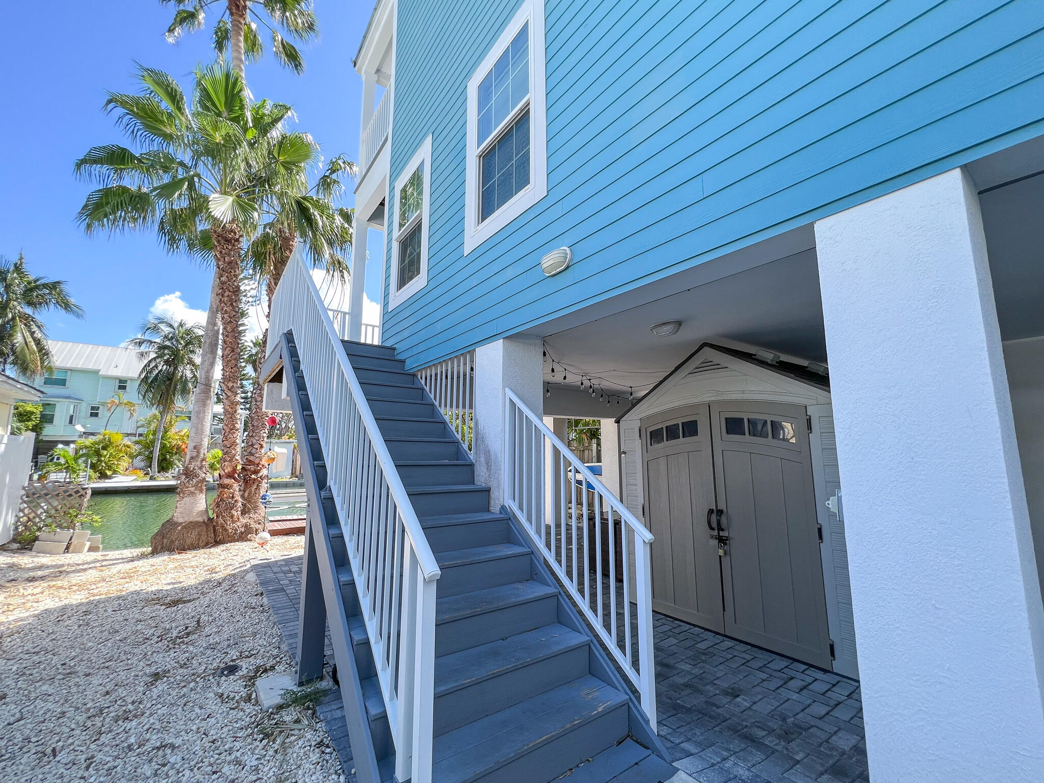 Blue raised coastal home with exterior stairs beside a palm-lined canal.