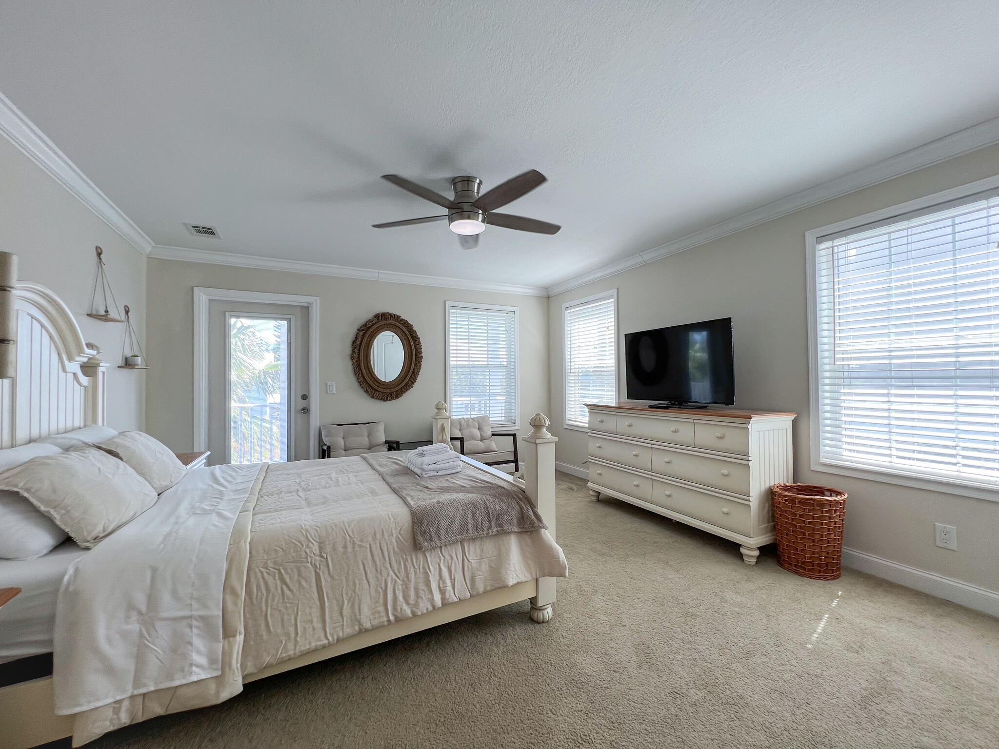 Furnished bedroom with a white bed, ceiling fan, television dresser, and windows with blinds.