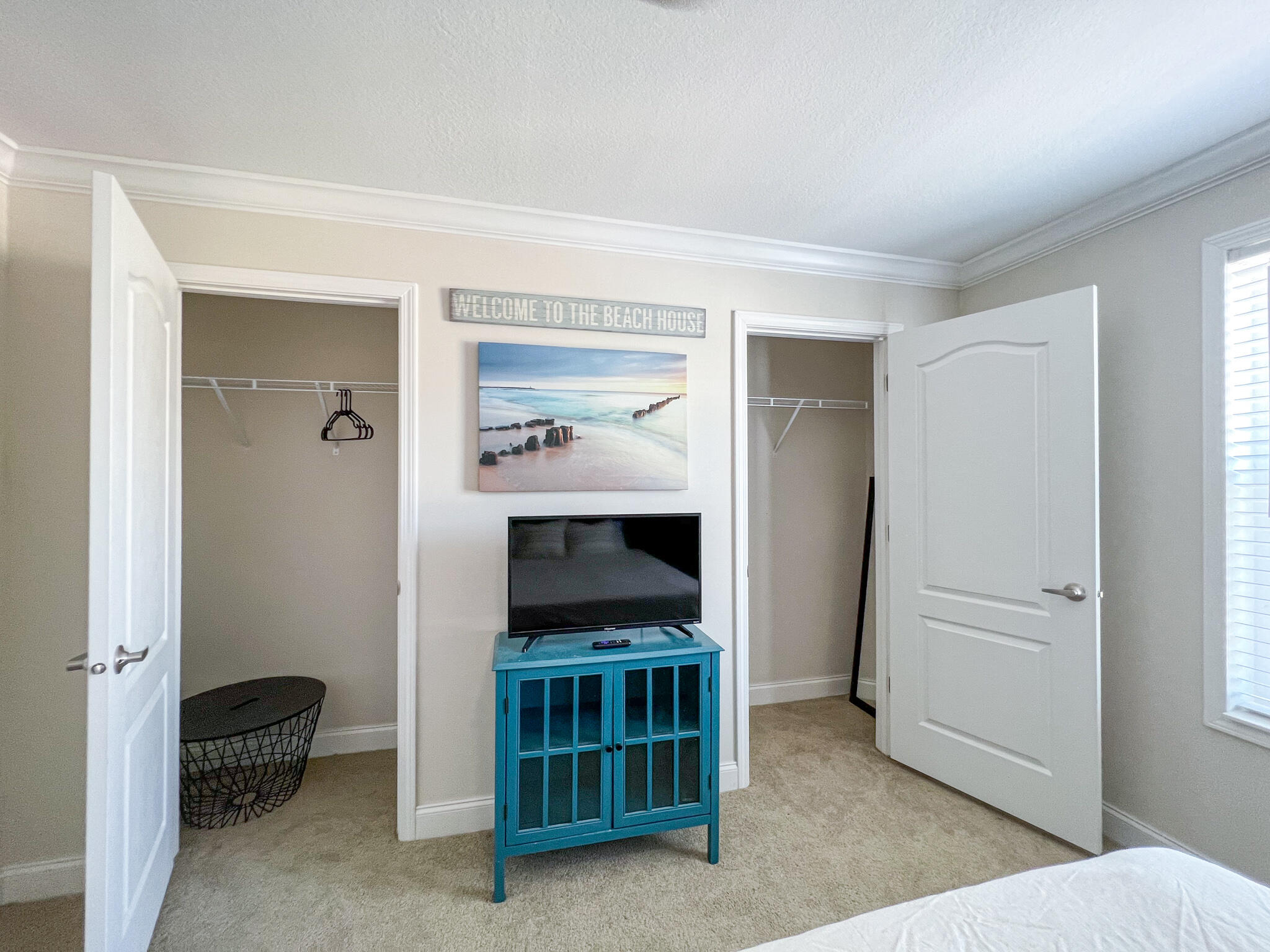 Bedroom with two open closets and a television on a turquoise cabinet beneath beach-themed wall art.