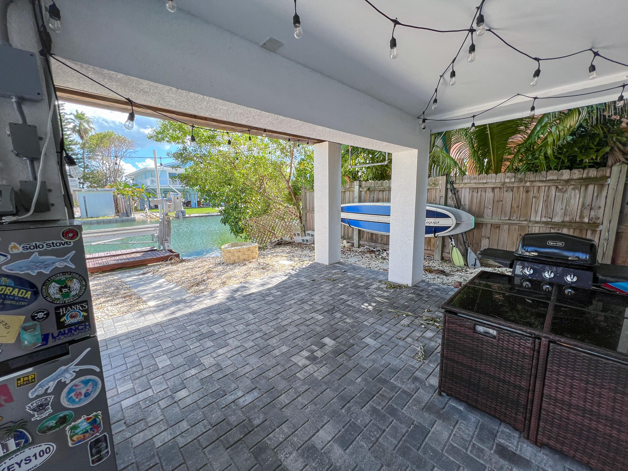 Covered canal-side patio with string lights, a grill, and a paddleboard beside the water.