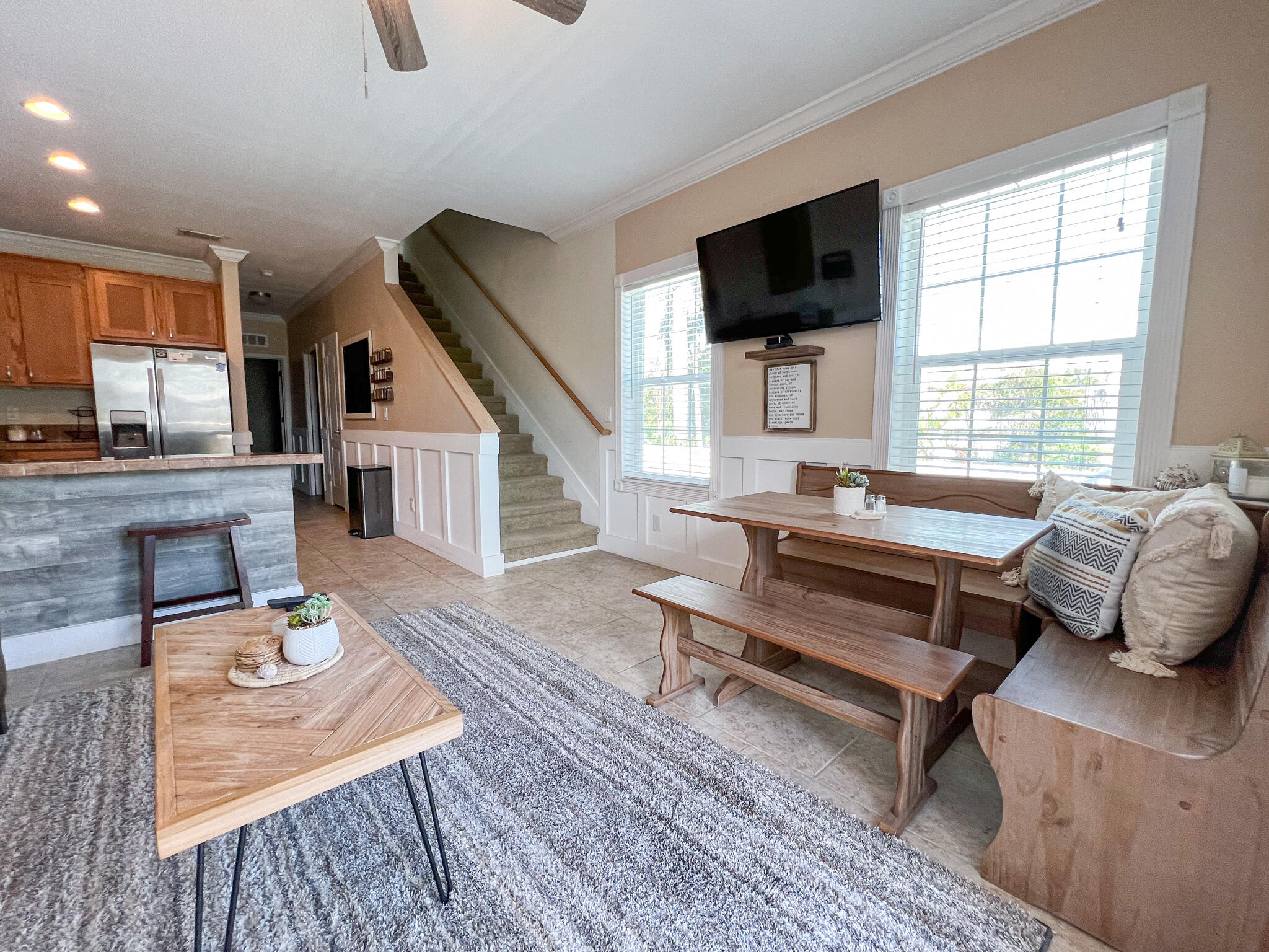 Open-plan living room with a kitchen bar, carpeted staircase, and wooden dining nook by large windows.