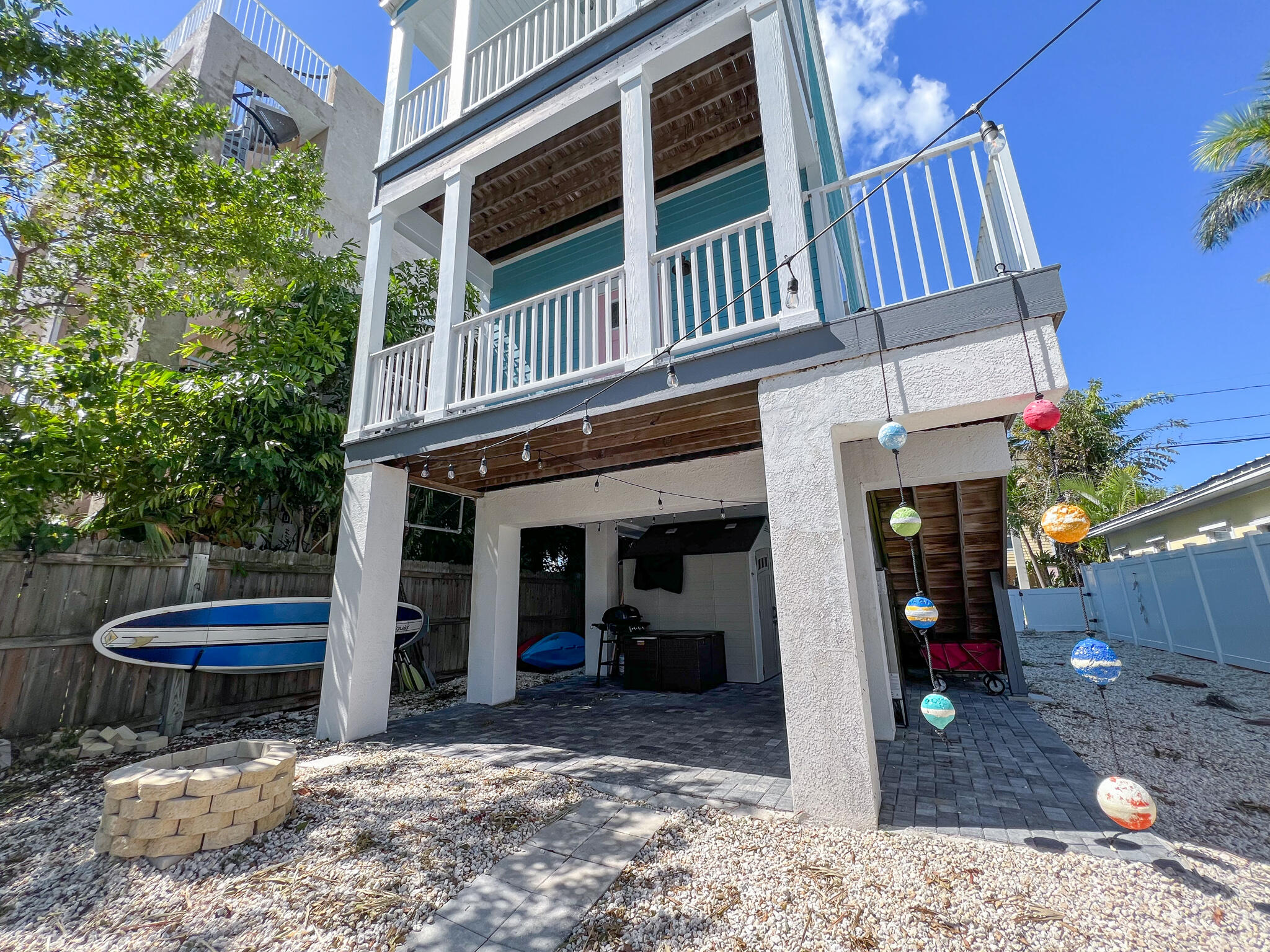 Raised coastal home with a covered patio, upper deck, paddleboards, and colorful hanging lights.