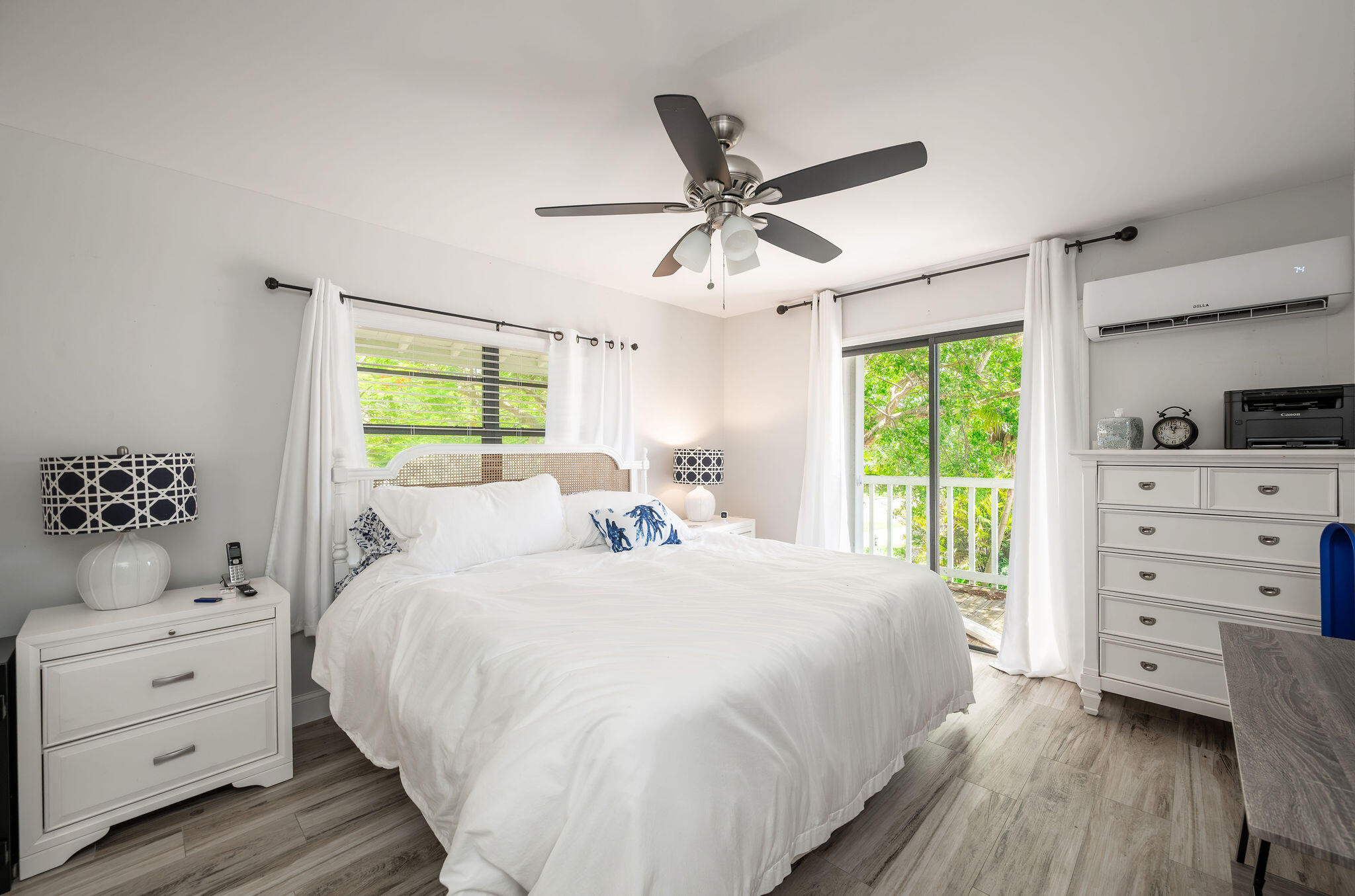 Bedroom with a white bed and dresser beside sliding doors opening onto a leafy balcony.