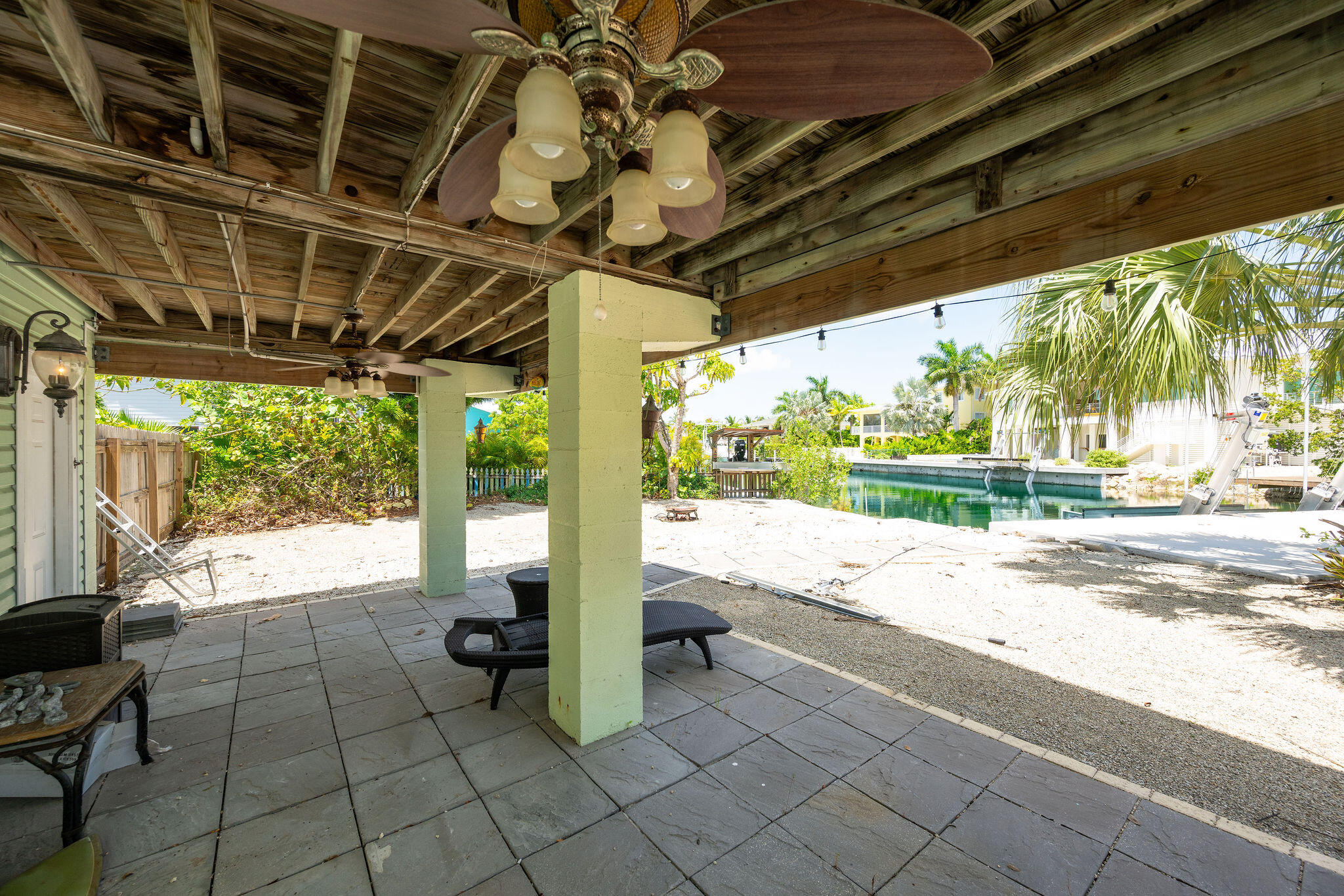 Covered patio overlooking a canal, with lounge furniture, palm trees, and boat lifts.