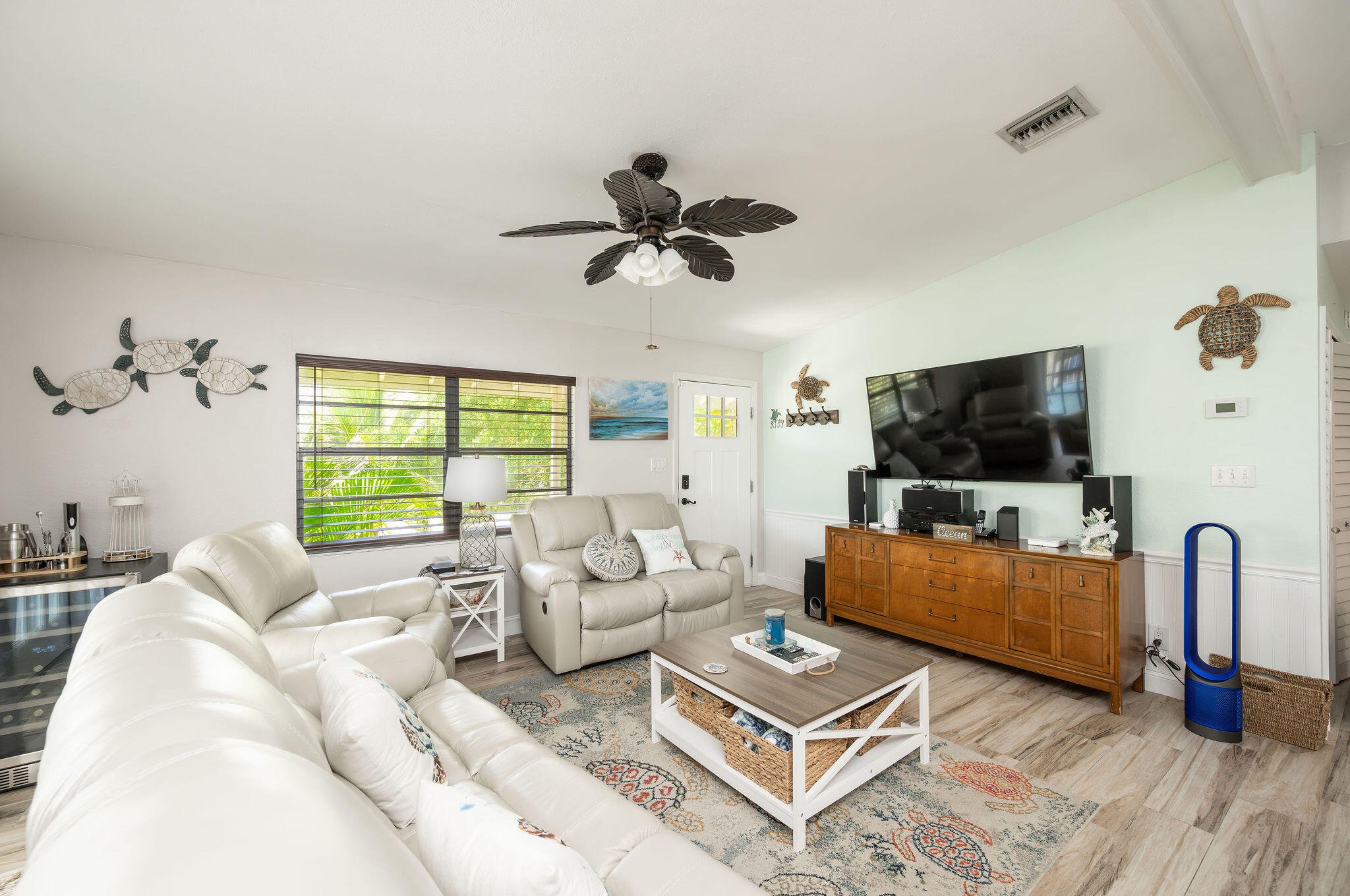 Furnished living room with white leather sofas, a large television, and a window overlooking tropical greenery.
