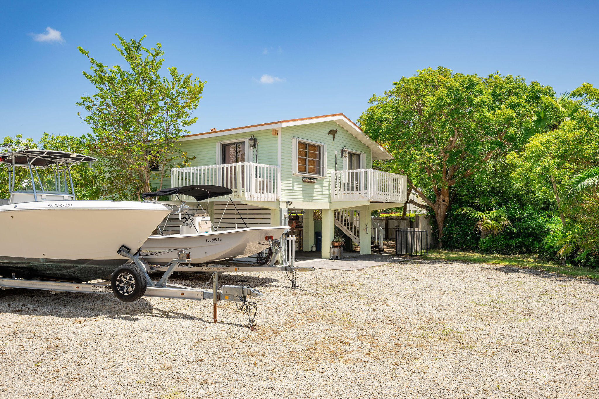 Elevated pale green home with white-railed balconies and two boats parked on a gravel lot.