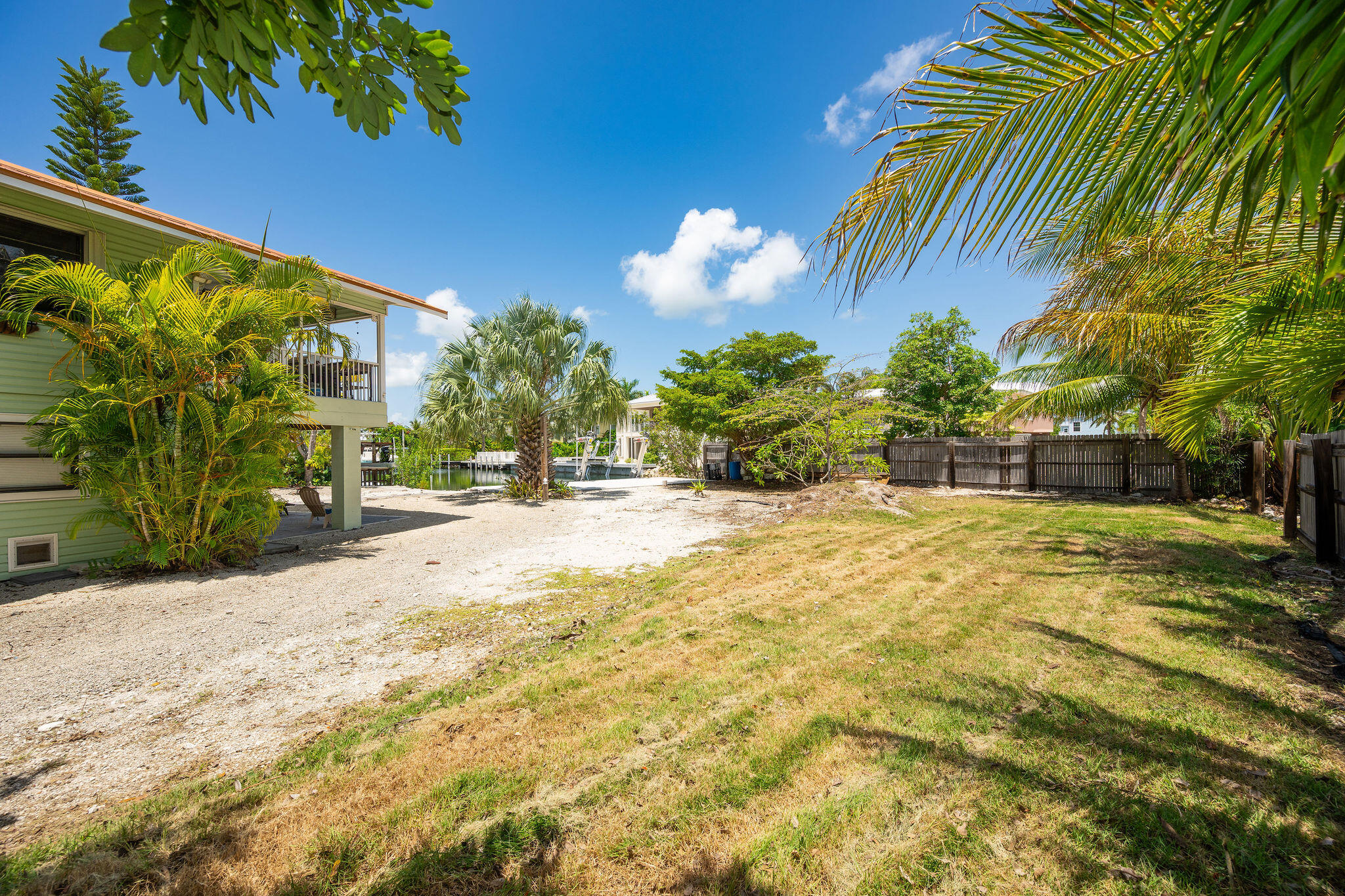 Backyard with a raised pale-green home, tropical palms, and a canal dock beyond the lawn.