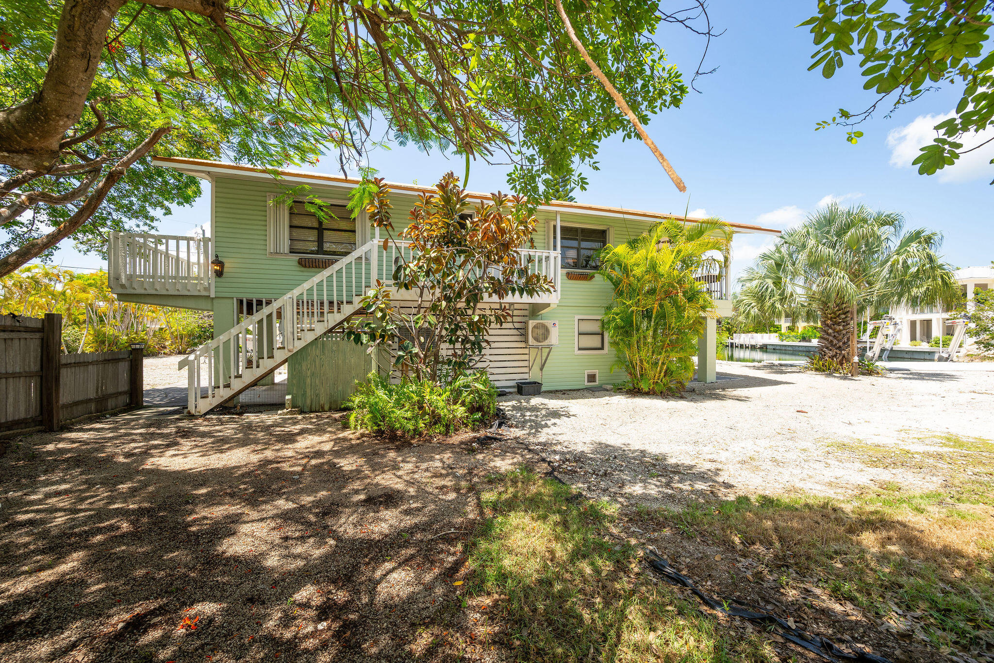 Pale green raised home with a white staircase and deck, tropical trees, and a canal nearby.