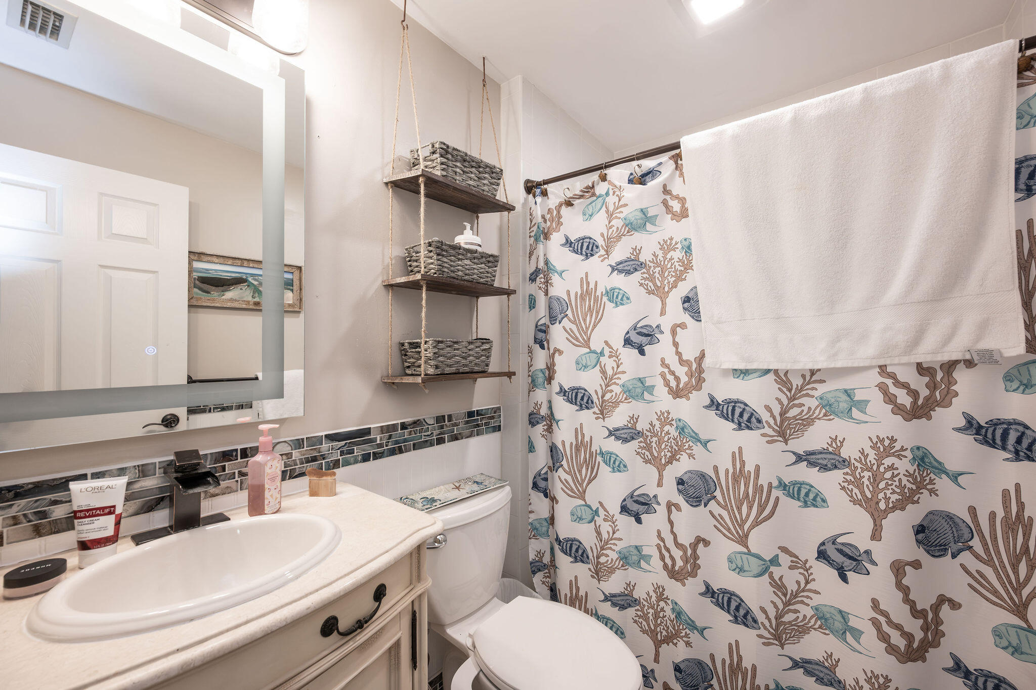 Bathroom with a cream vanity, fish-patterned shower curtain, and rope-hung basket shelves.