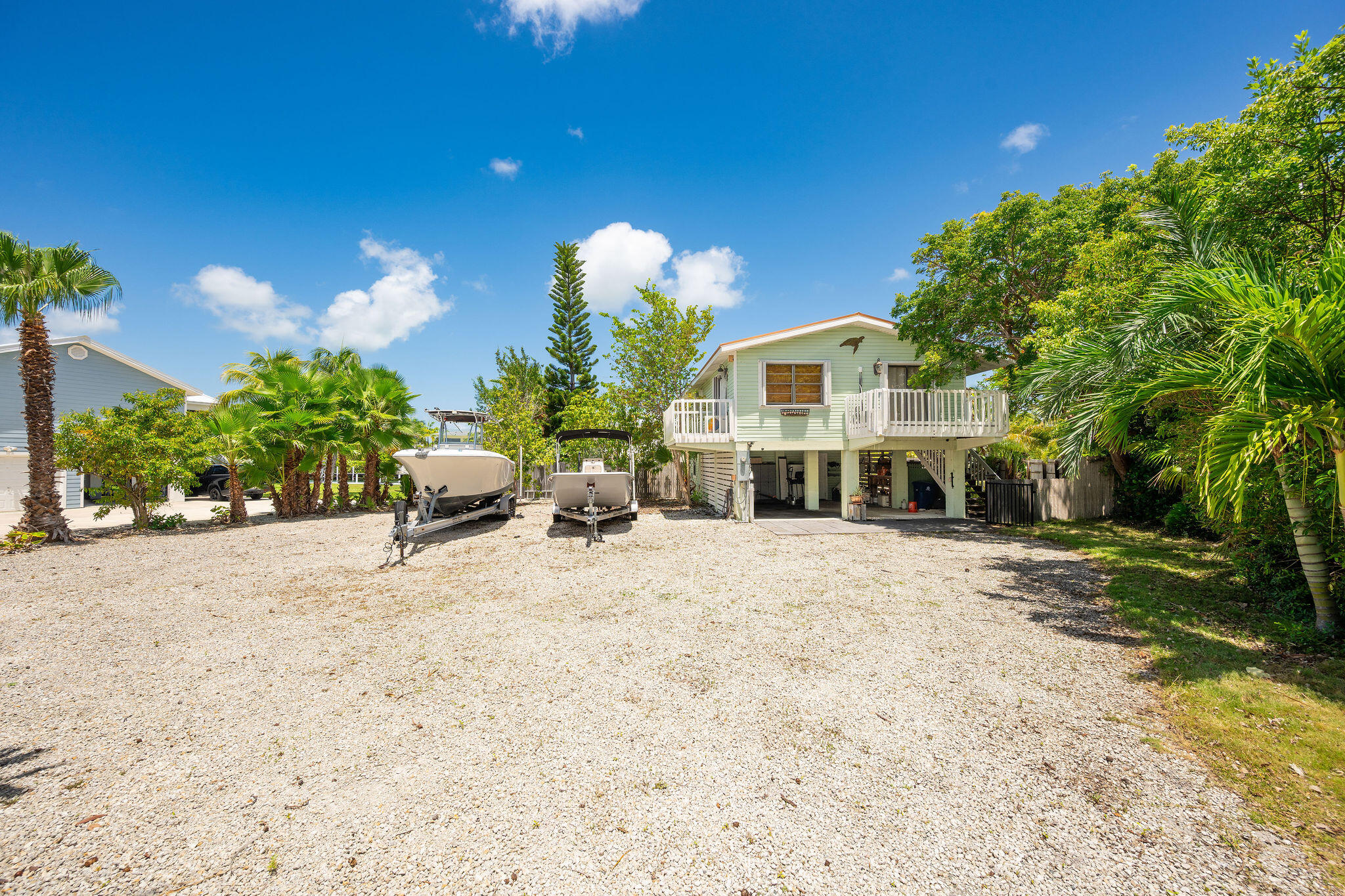 Raised light green home with a white deck, two boats on trailers, and tropical landscaping in a gravel yard.