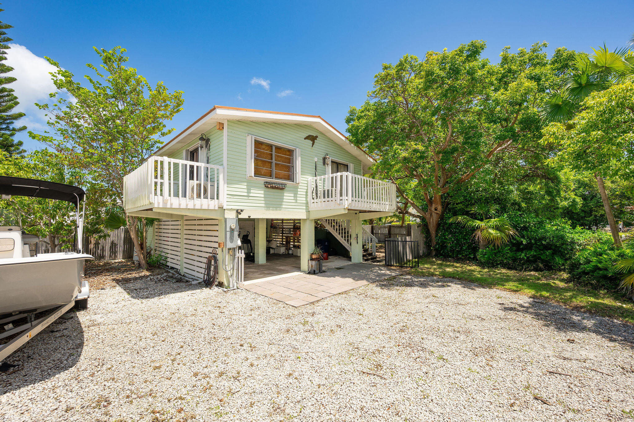 Elevated mint-green home with white balconies, a gravel yard, and a boat beneath leafy trees.