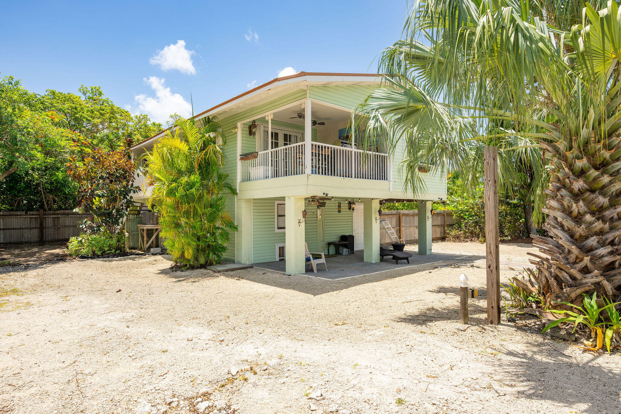 Raised pale green house with a covered balcony and patio, framed by palm trees in a sandy yard.