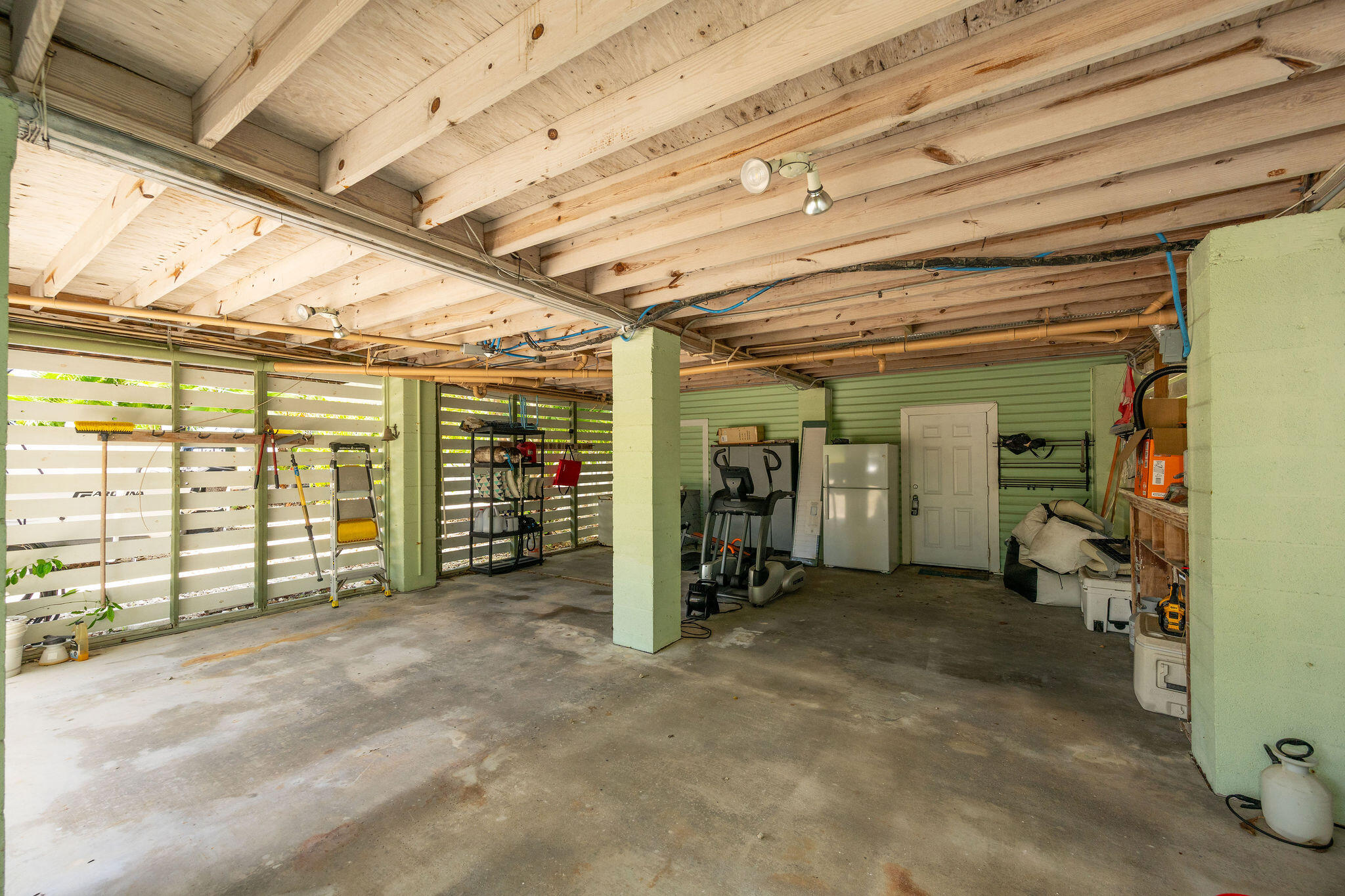 Garage with exposed wood beams, slatted walls, exercise equipment, and a refrigerator.