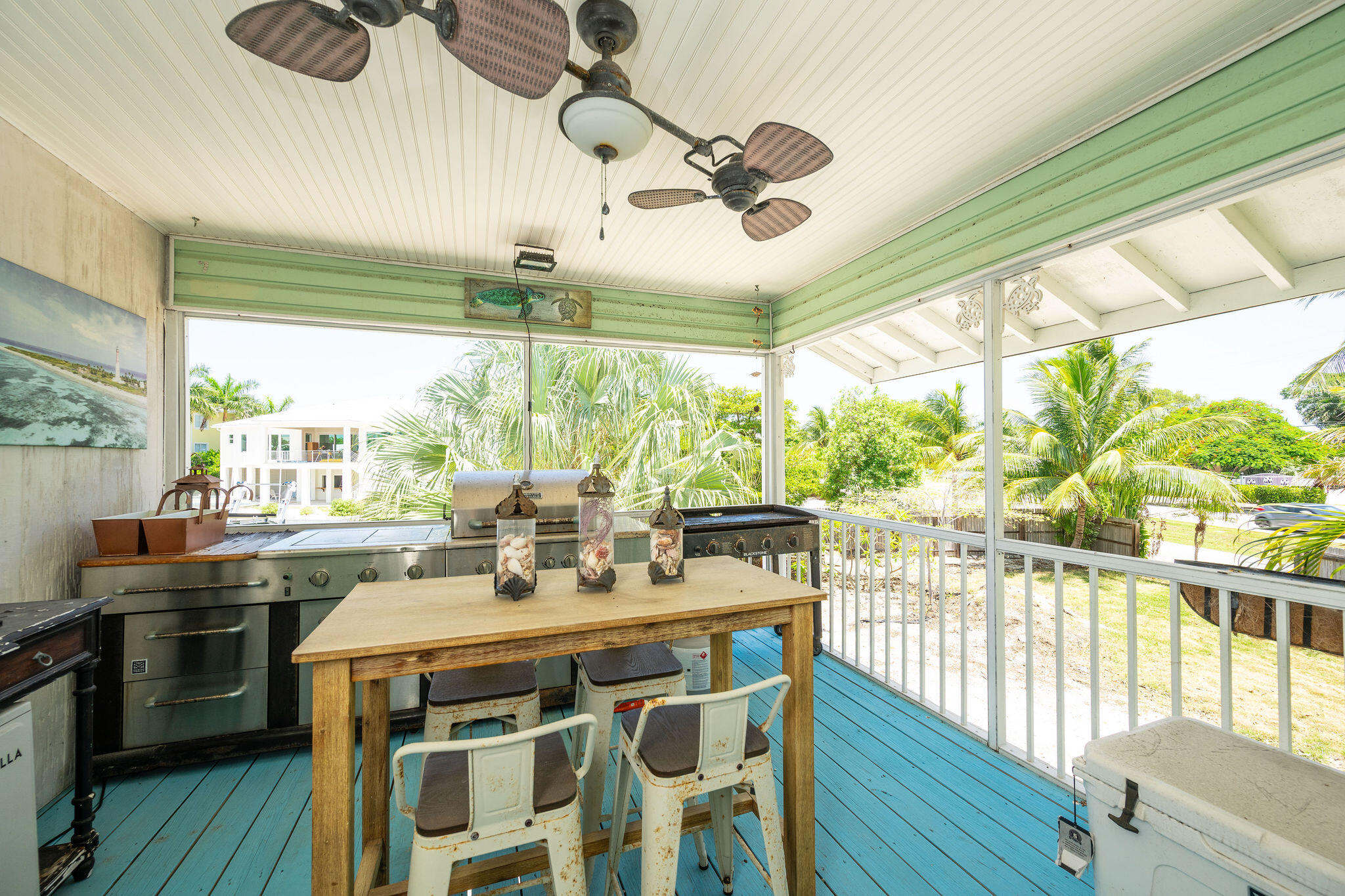 Covered porch outdoor kitchen with a stainless steel grill, wooden dining table, and palm trees beyond the railing.