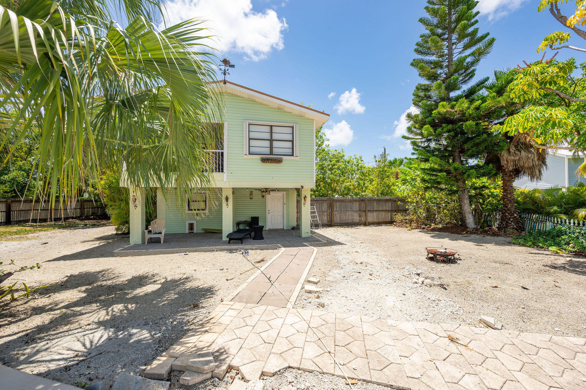 Raised pale green house with a shaded patio and tropical trees in the gravel yard.