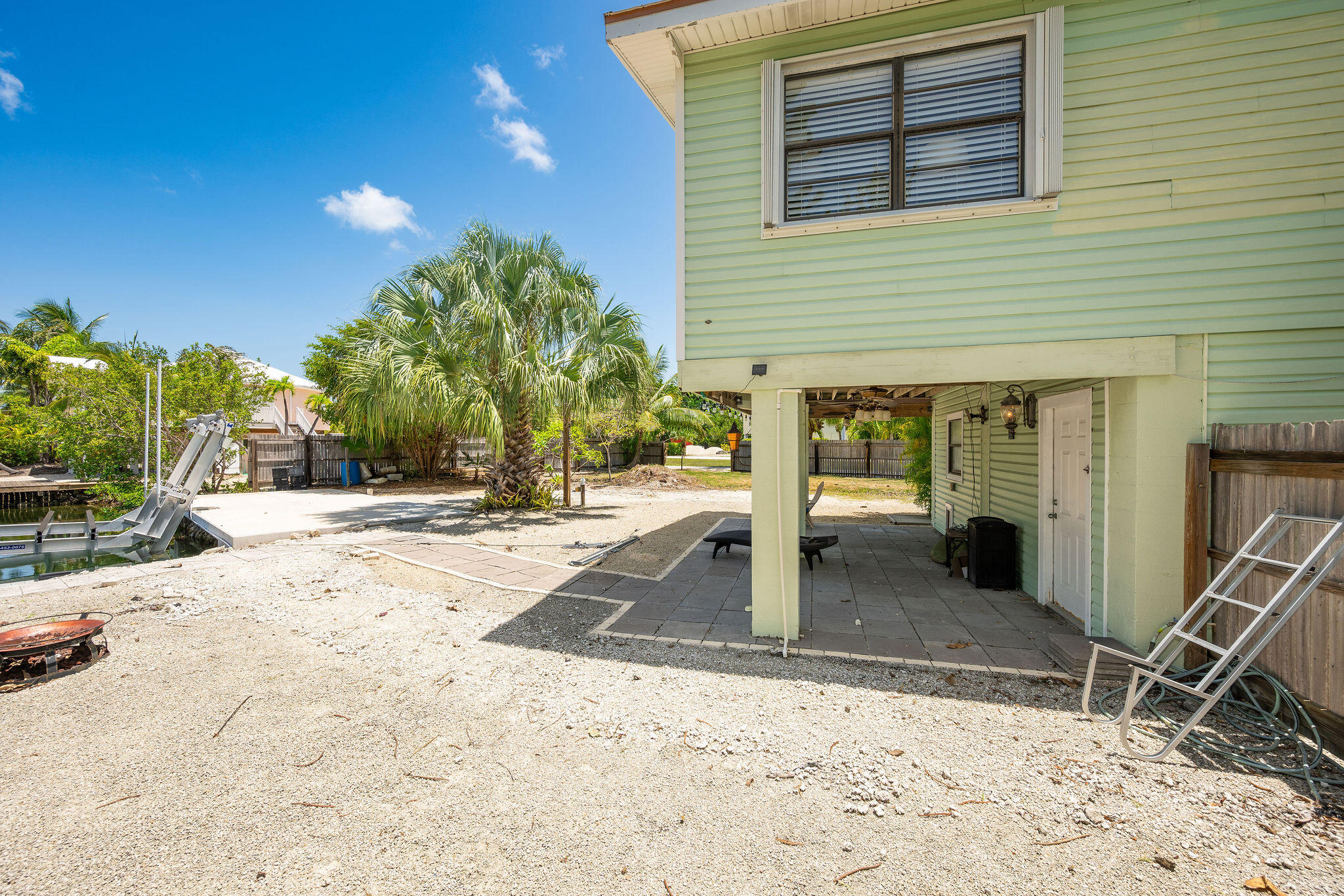 Raised light-green home with a covered patio, palm trees, gravel yard, and canal boat lift.