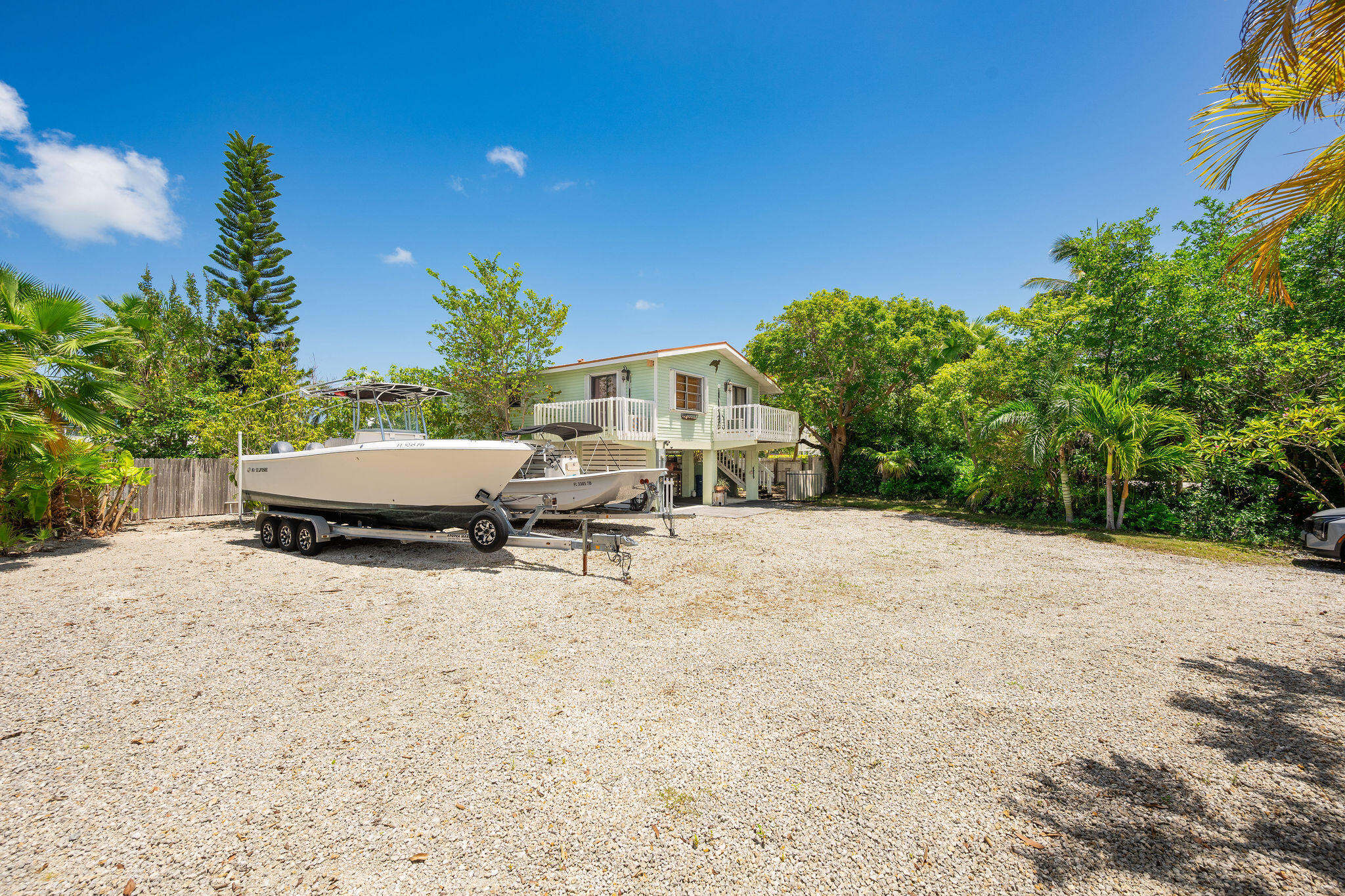 Elevated pale-green home with a white balcony and two boats parked in its gravel yard