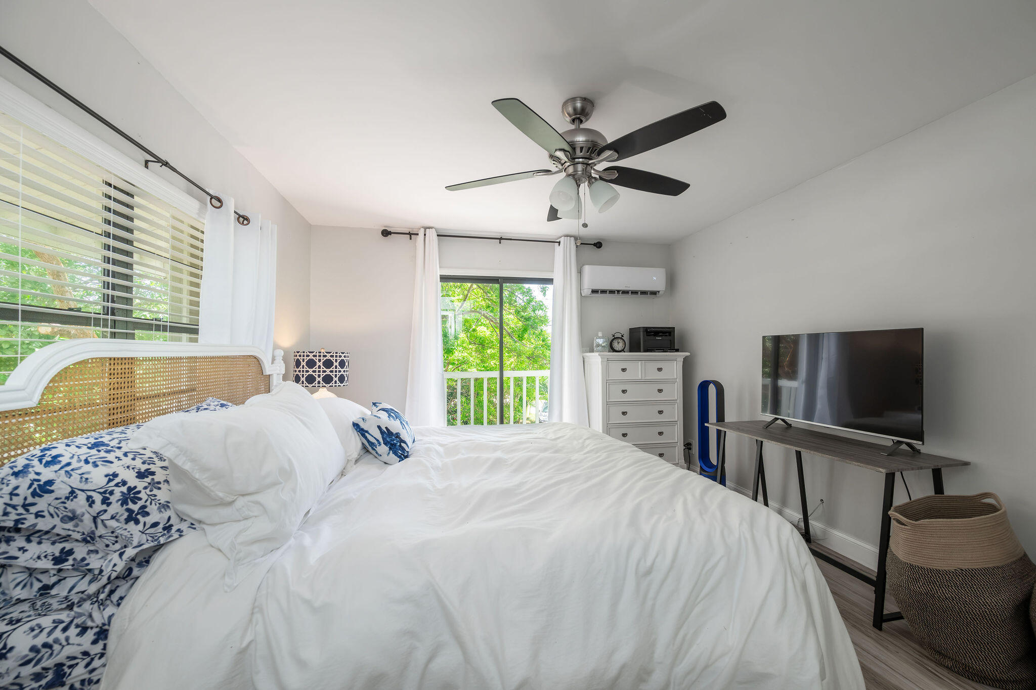 Furnished bedroom with a white bed, woven headboard, blue patterned pillows, and leafy views through sliding doors.
