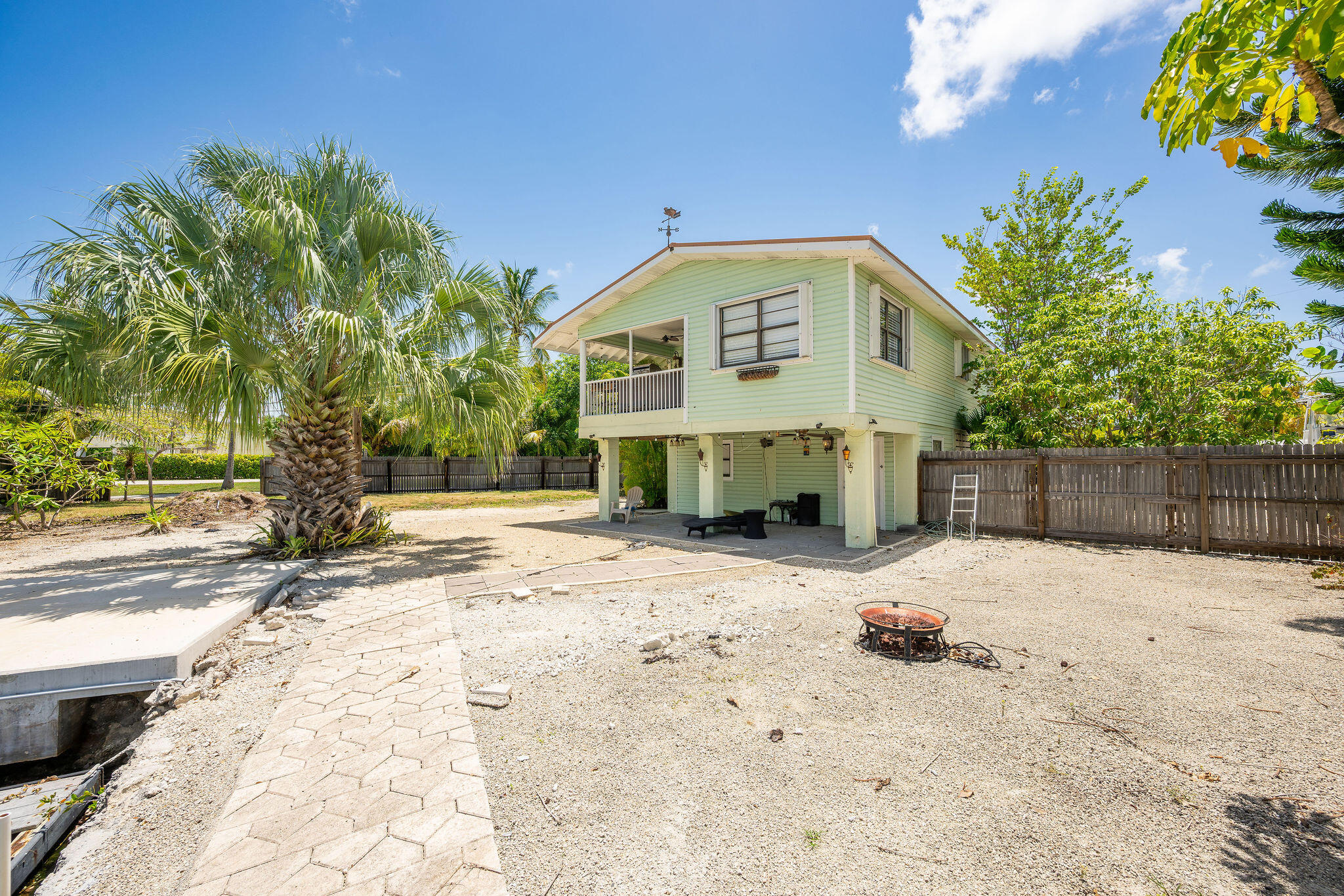 Raised pale green house with a covered balcony, gravel yard, paver path, and palm trees.