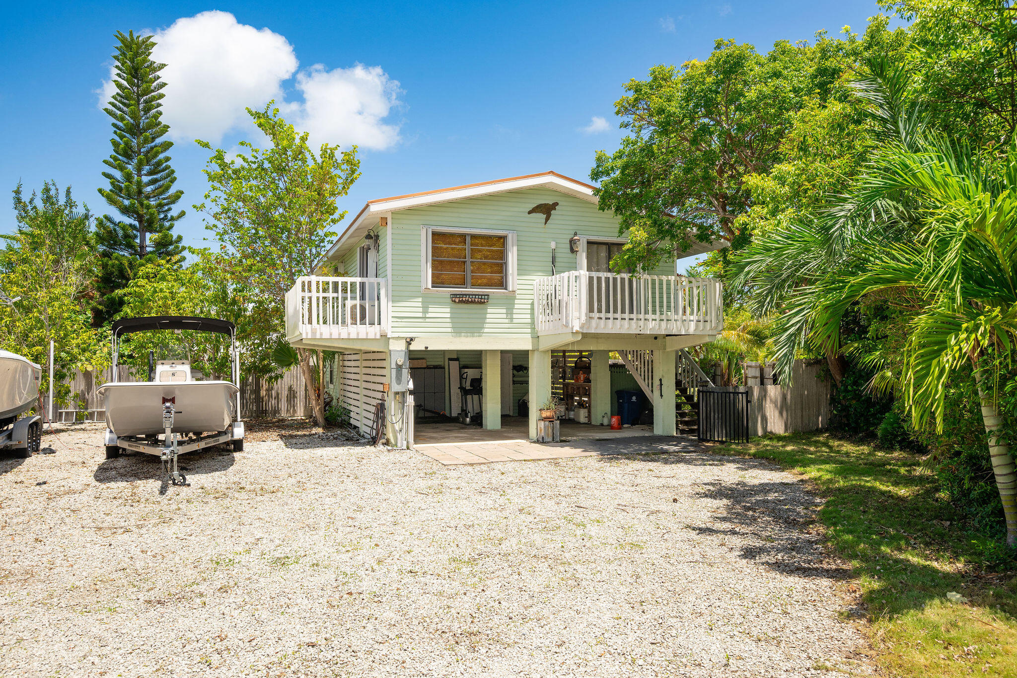 Elevated pale green home with white decks, a boat on a trailer, and tropical landscaping.