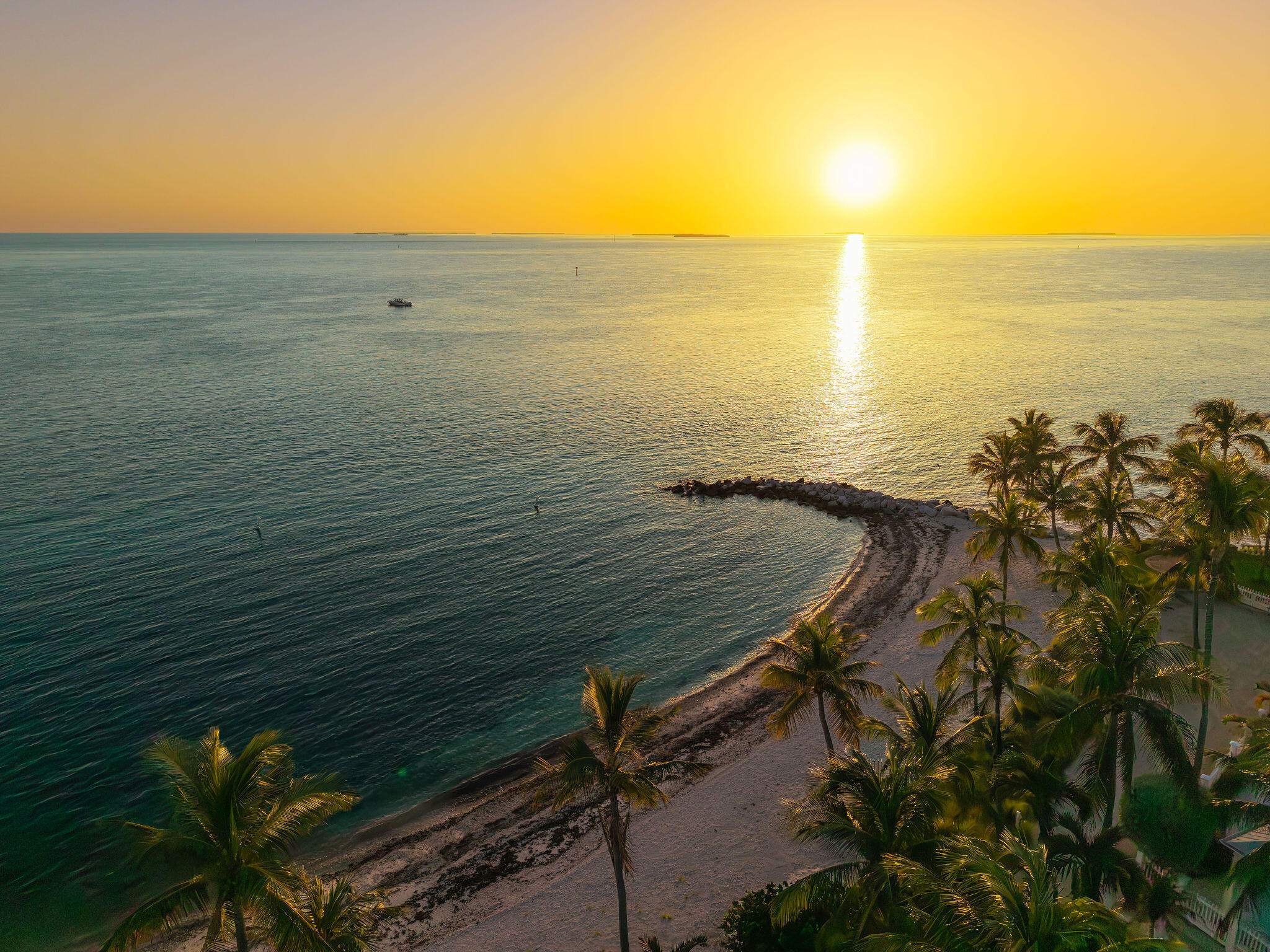 Palm-lined beach and turquoise water at sunset, with a rocky breakwater and sunlit sea.