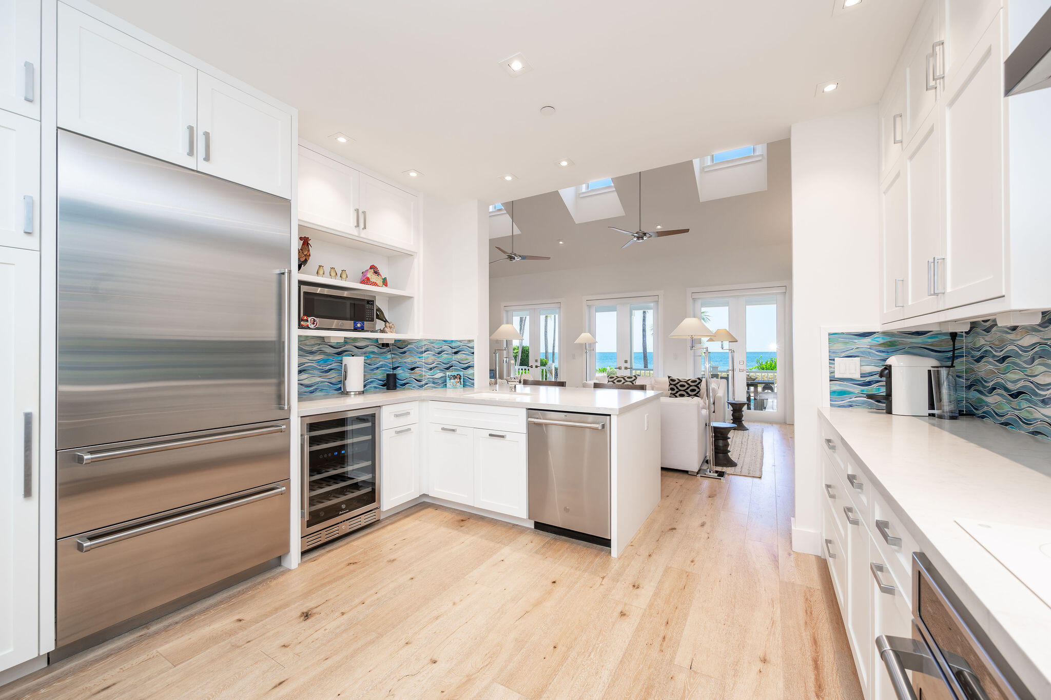 Bright white kitchen with stainless appliances and blue wave backsplash, opening toward an ocean view.