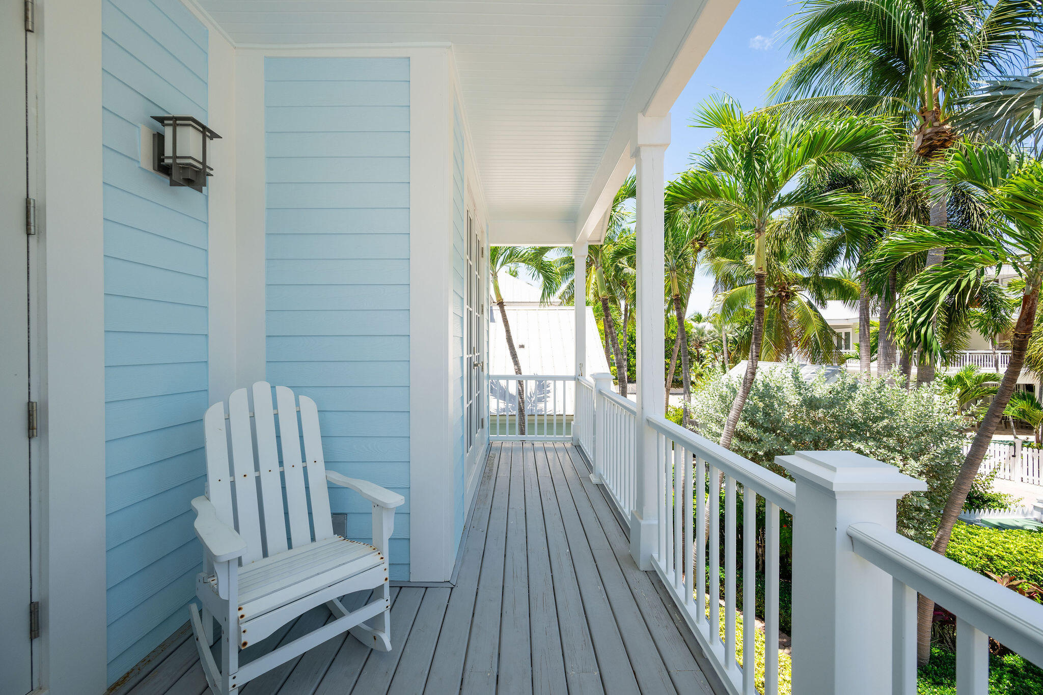 Covered porch with a white Adirondack chair, gray plank floor, and views of palm trees.