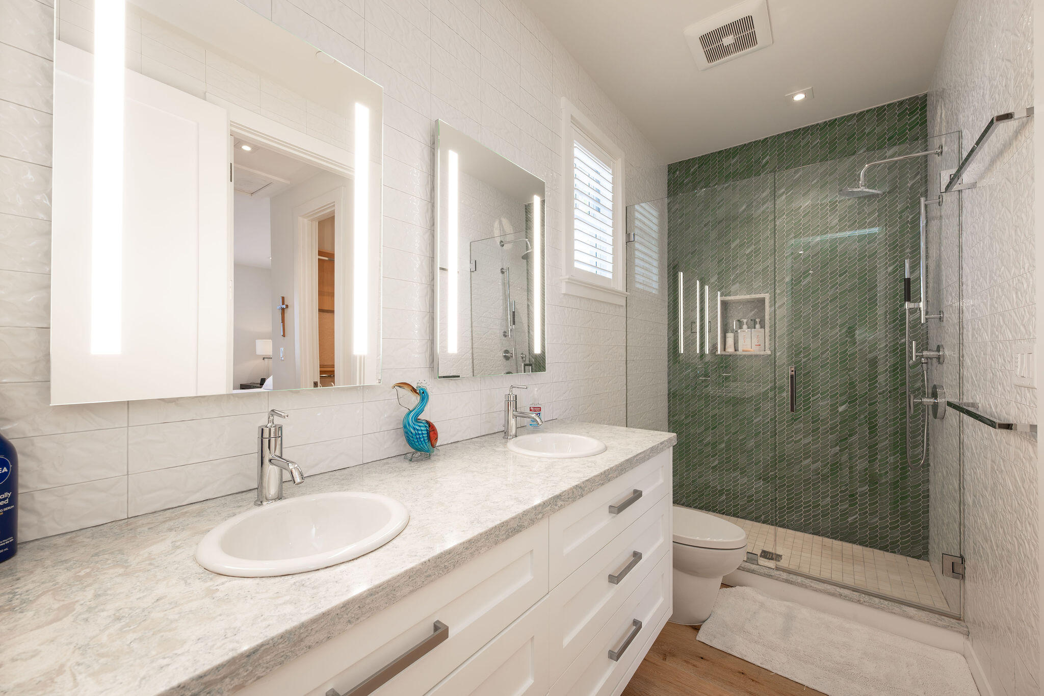 Bathroom with a double-sink vanity and glass shower backed by dark green tile.