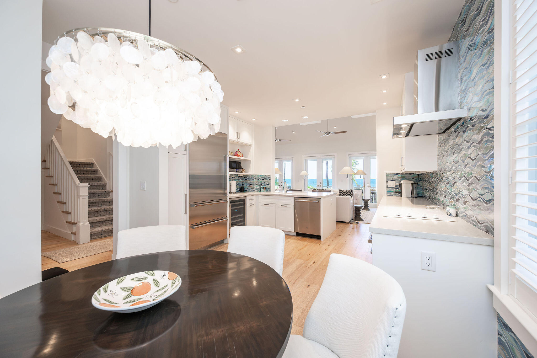 Open-plan kitchen with blue wave-patterned tile, a large chandelier, and ocean views through the rear doors.