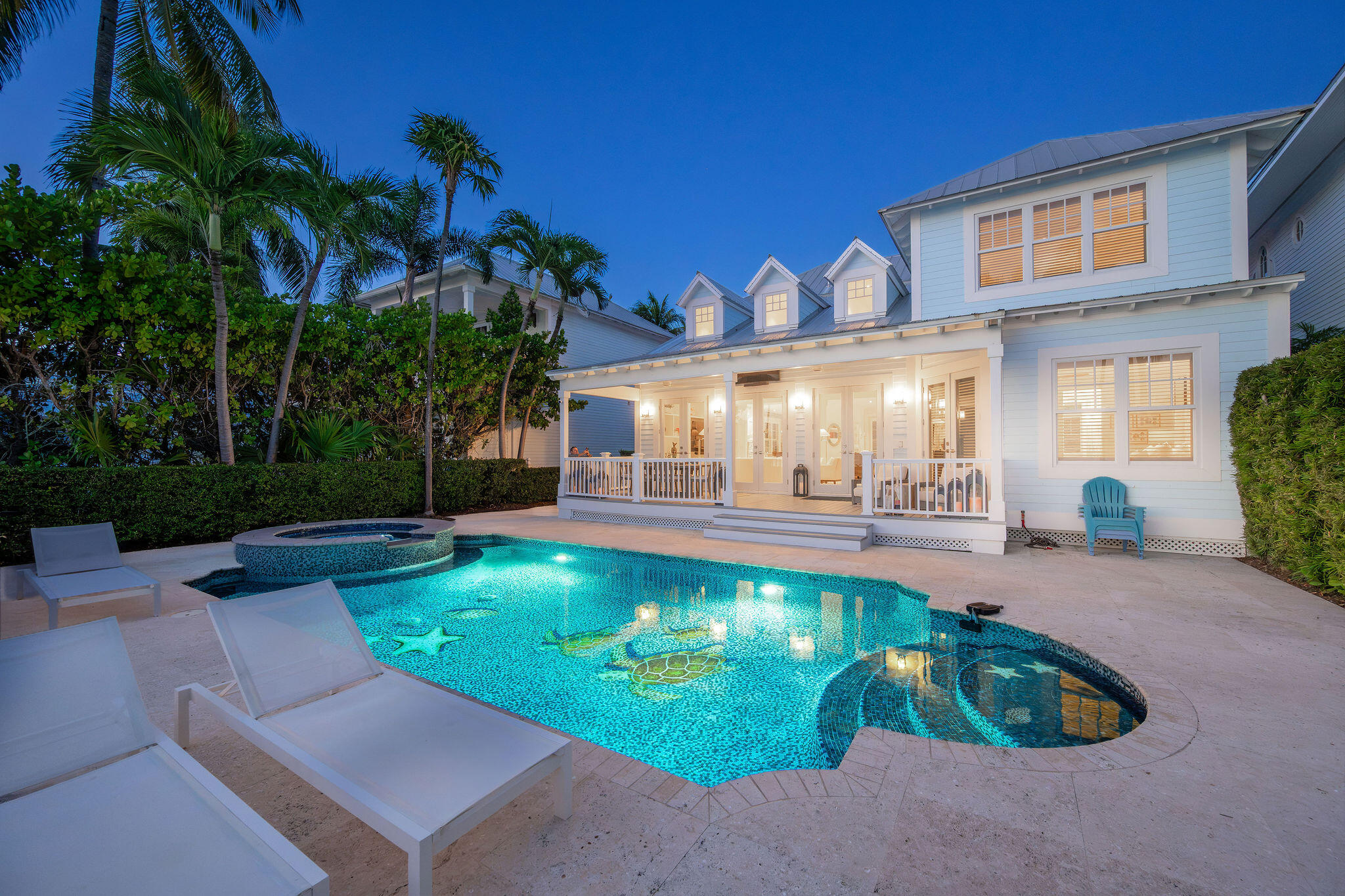 White coastal home glowing at dusk behind a turquoise pool and round spa, framed by palm trees.