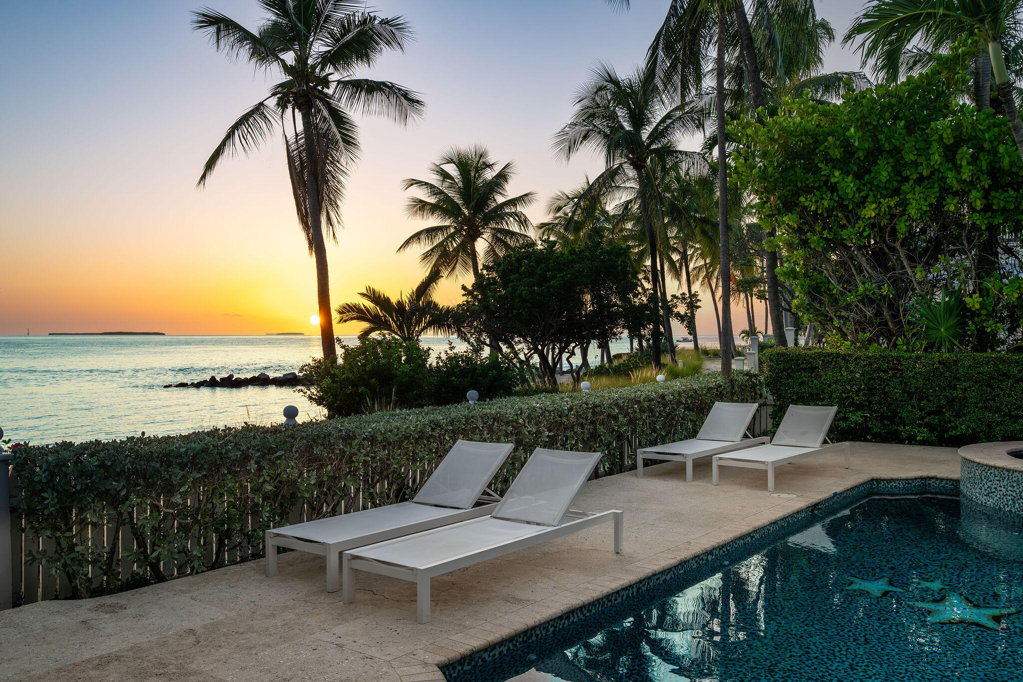 Waterfront pool terrace with lounge chairs and palm trees overlooking the ocean at sunset.