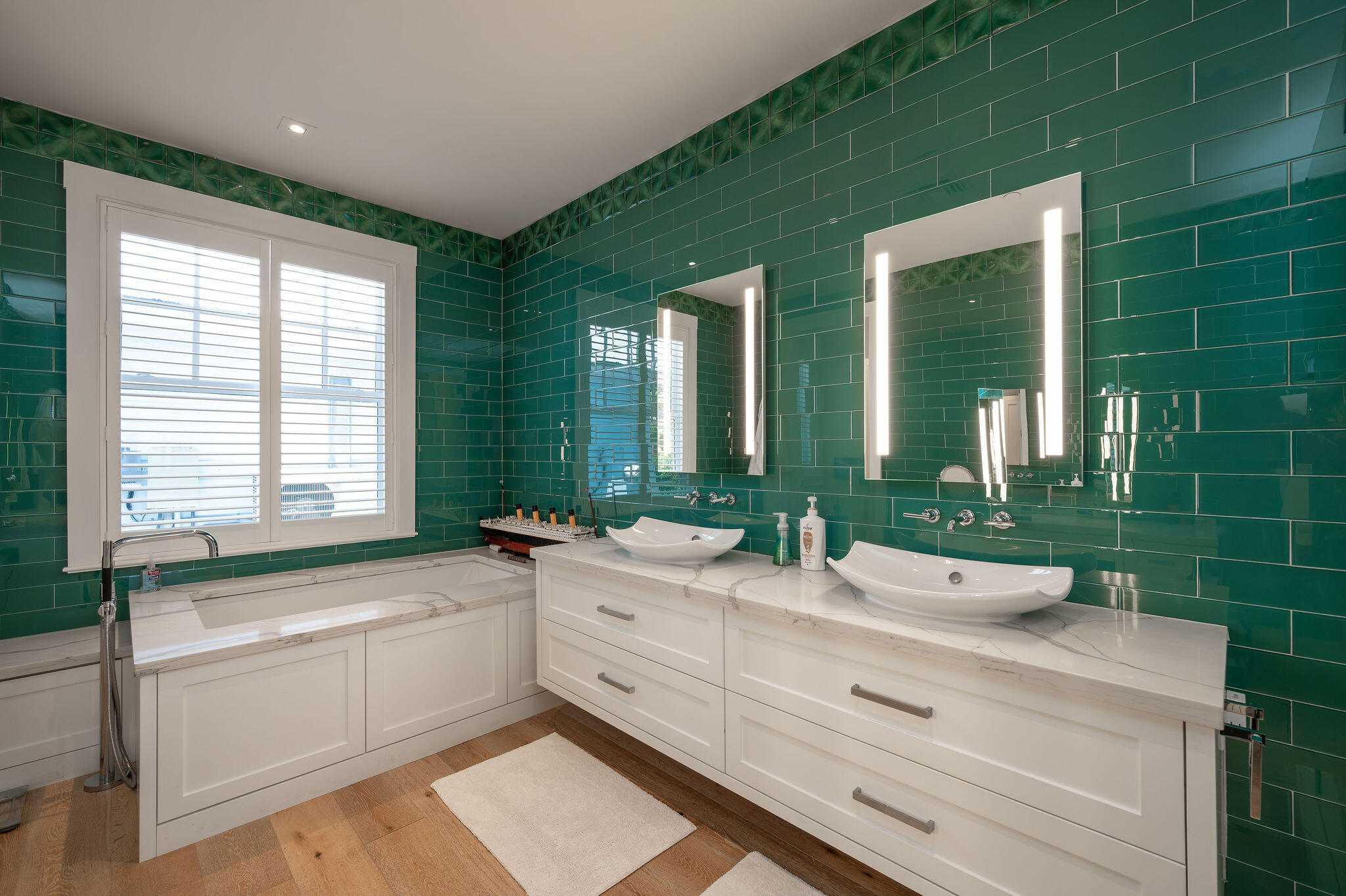 Modern bathroom with emerald tile, double vanity, illuminated mirrors, and a soaking tub beneath shuttered windows.